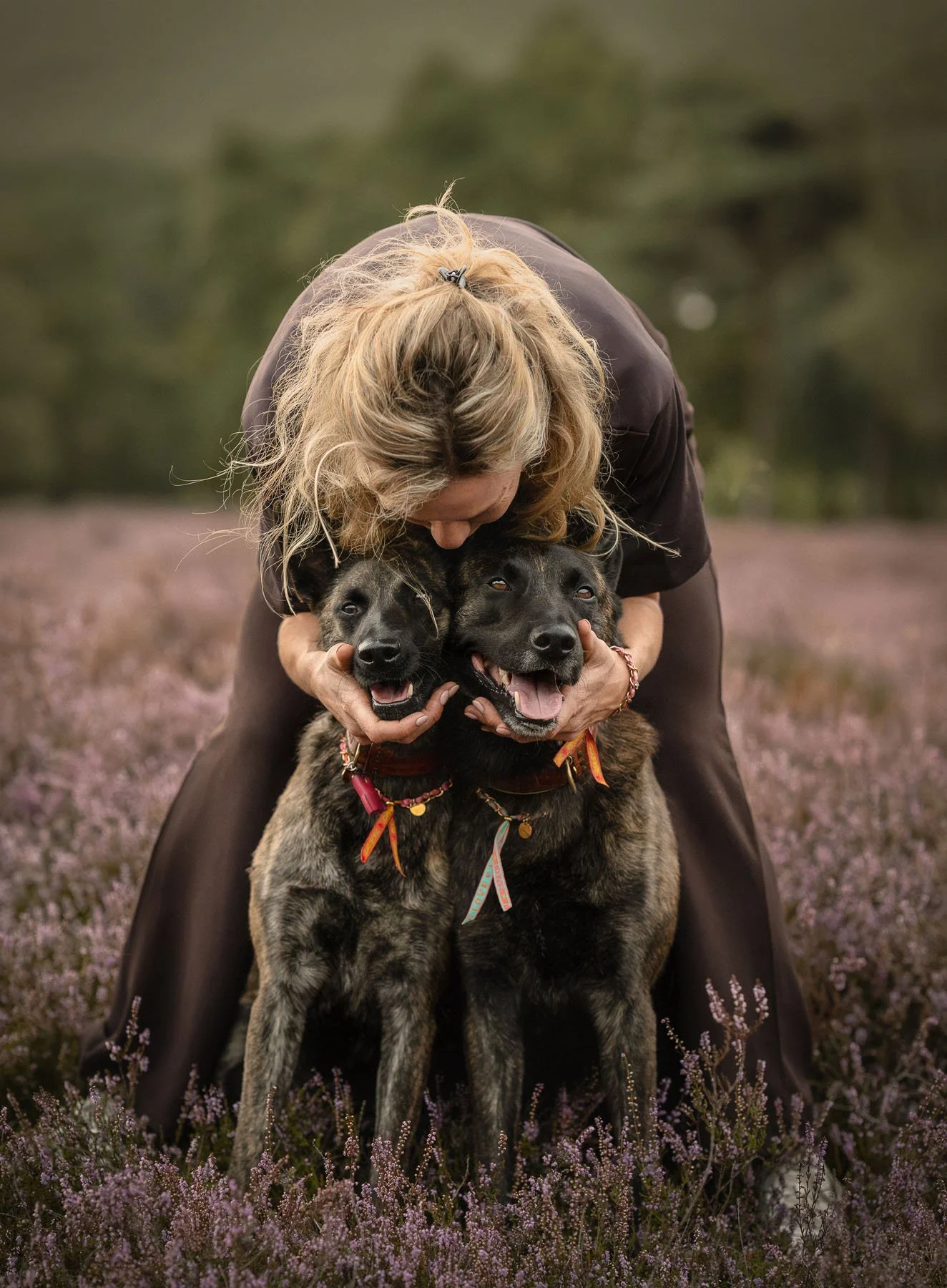 Een vrouw omarmt twee honden in een veld met paarse bloemen, met een groene achtergrond.
