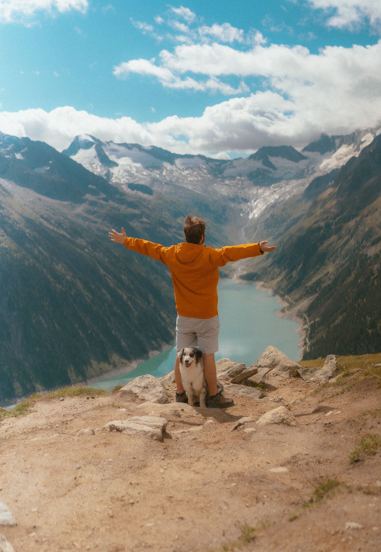 Man in oranje jas met armen wijd gestrekt, naast een hond, op een berg met uitzicht op een fjord en besneeuwde bergtoppen onder een blauwe hemel met wolken.