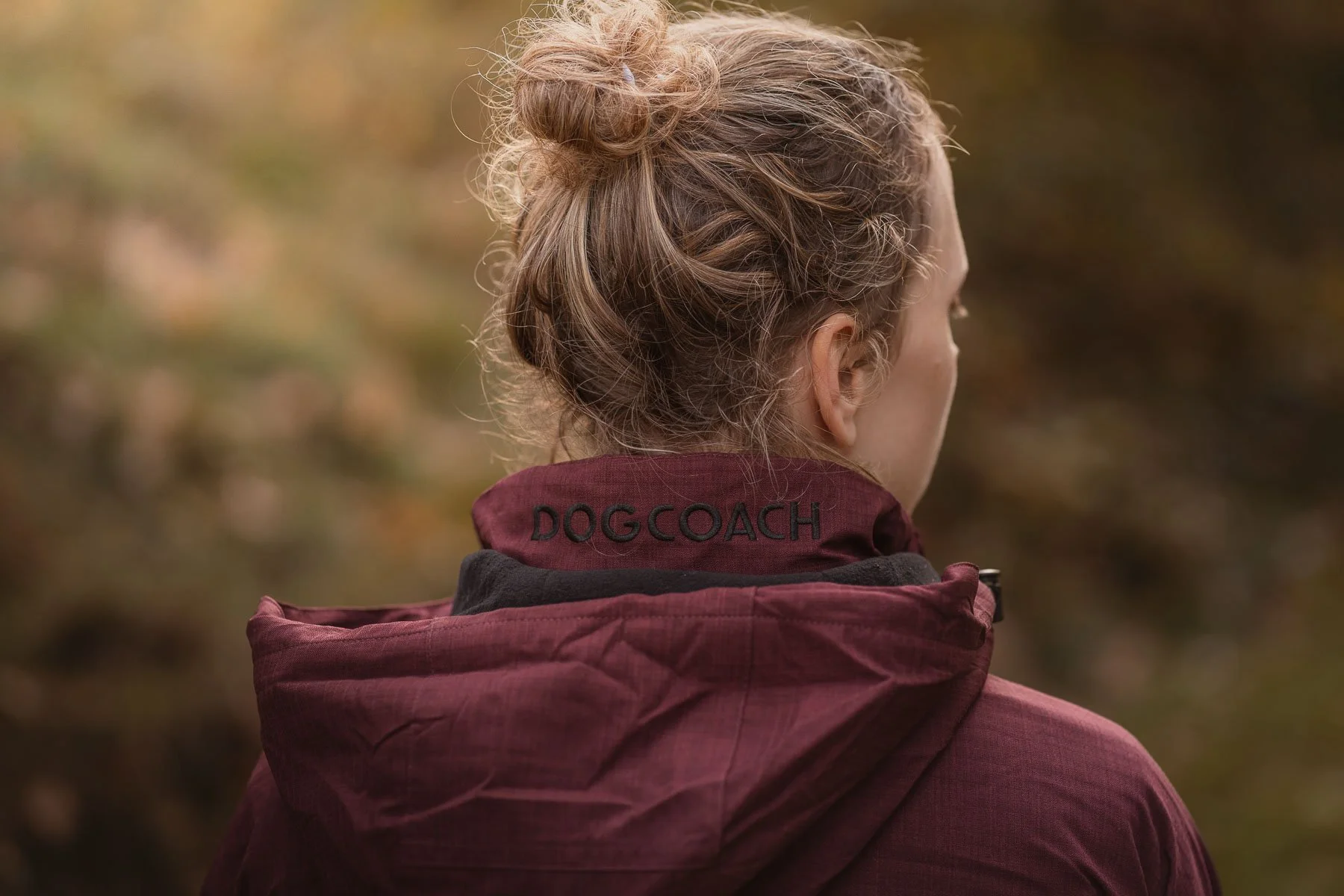 Een vrouw kijkt weg, draagt een rode jas met de tekst 'DOGCOACH' op de kraag, in een bosrijke omgeving met herfstbladeren.