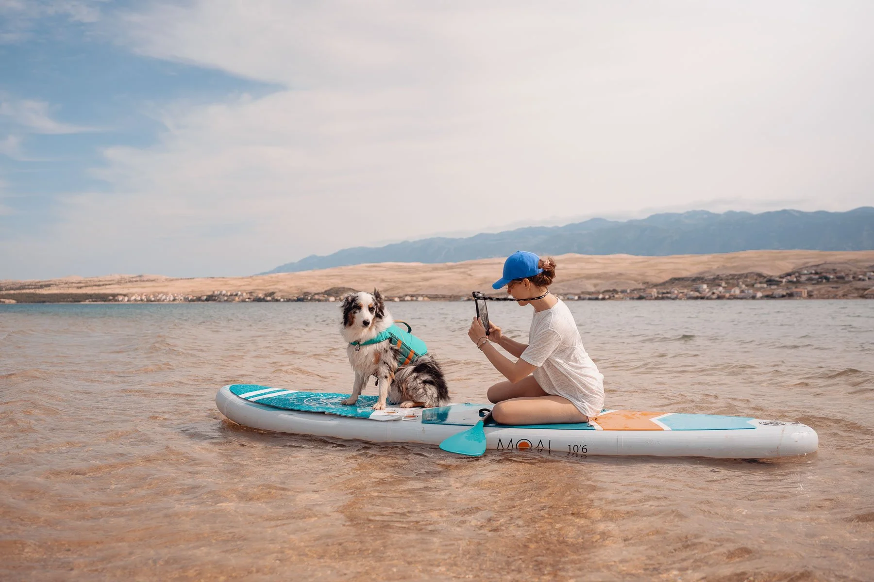 Een vrouw zit op een paddleboard in het water en maakt een foto van een border collie die op het paddleboard staat, met een landschap en bergen op de achtergrond.