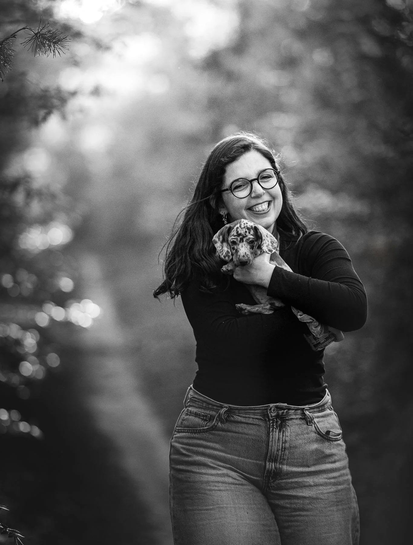 Een lachende vrouw met bruin haar en glasses houdt een speelse puppy vast. De achtergrond is wazig met bokeh en groene tinten.