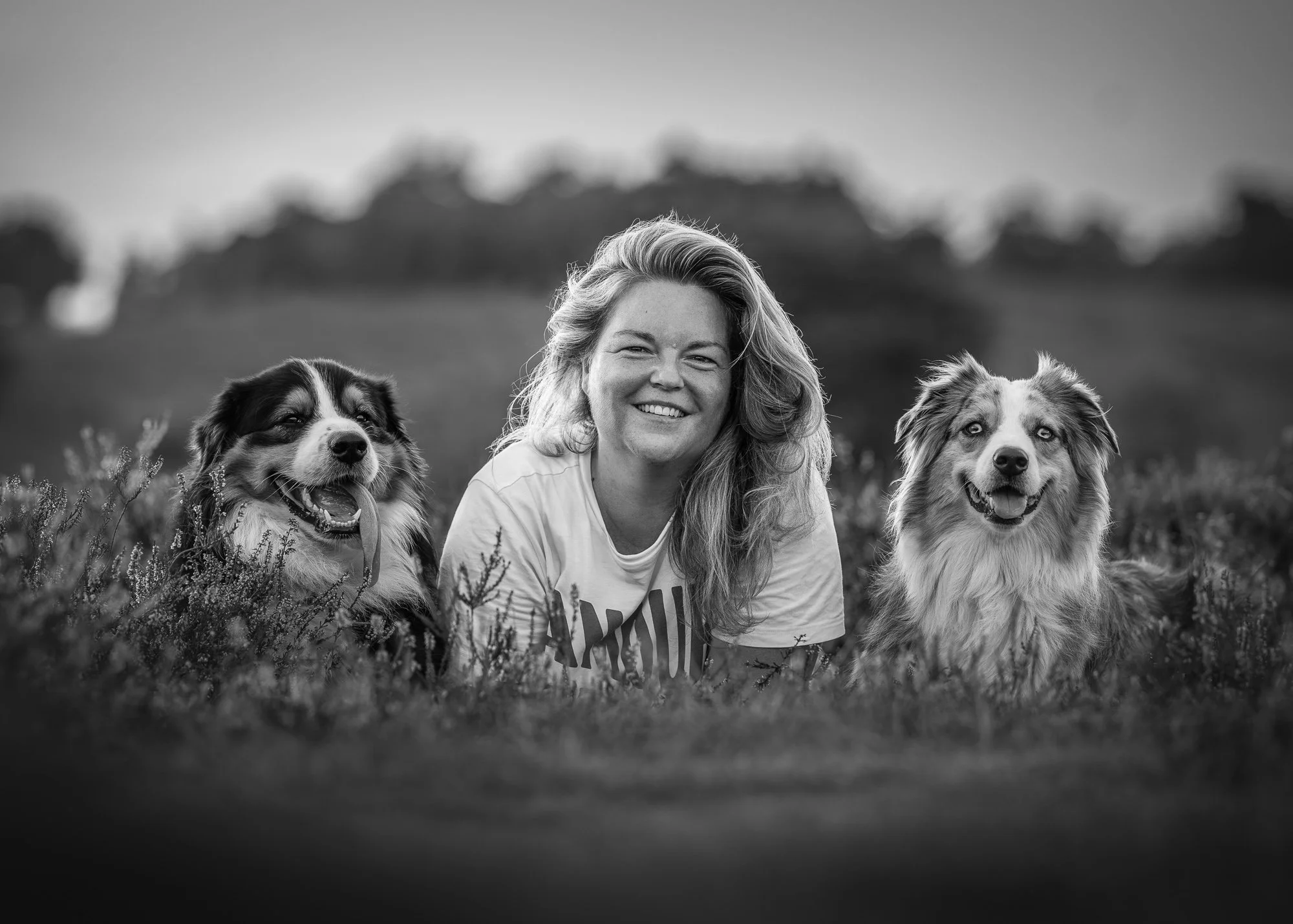 Zwart-witfoto van een vrouw die buiten op het gras ligt en glimlacht, met aan beide kanten van haar twee honden; op de achtergrond een landschap met bomen.