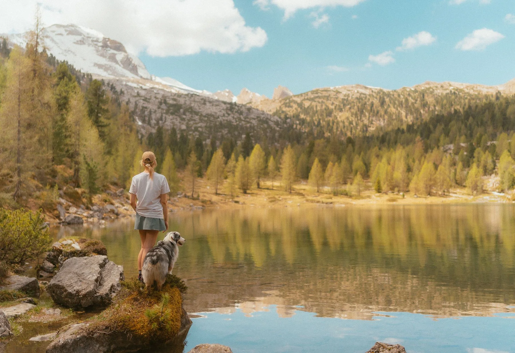 Een persoon en een hond staan aan de rand van een rustig meer, omringd door bergen en dennenbomen, onder een blauwe lucht met wolken.