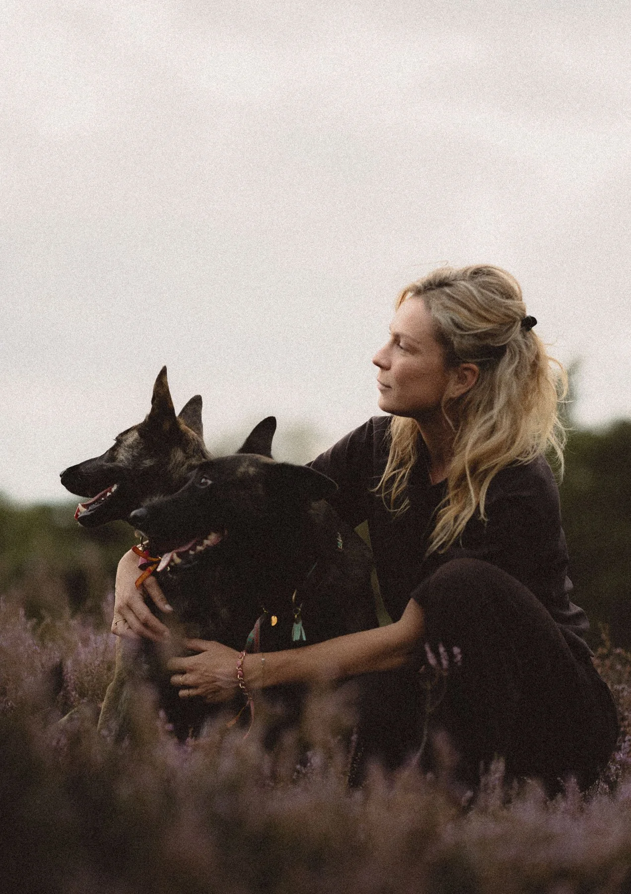 Vrouw die op haar knieën zit in een veld met bloemen, omarmt twee honden en kijkt naar de horizon tijdens zonsondergang.