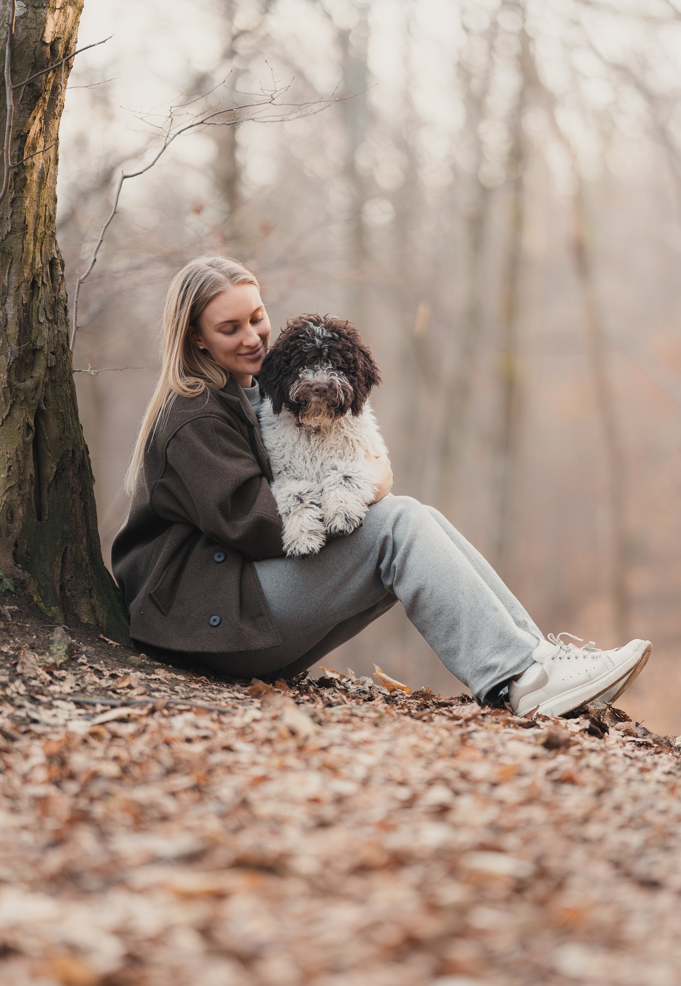 Een vrouw zit tegen een boom in een bos met gevallen bladeren en houdt een zwart-witte pluizige hond vast tijdens de herfst.