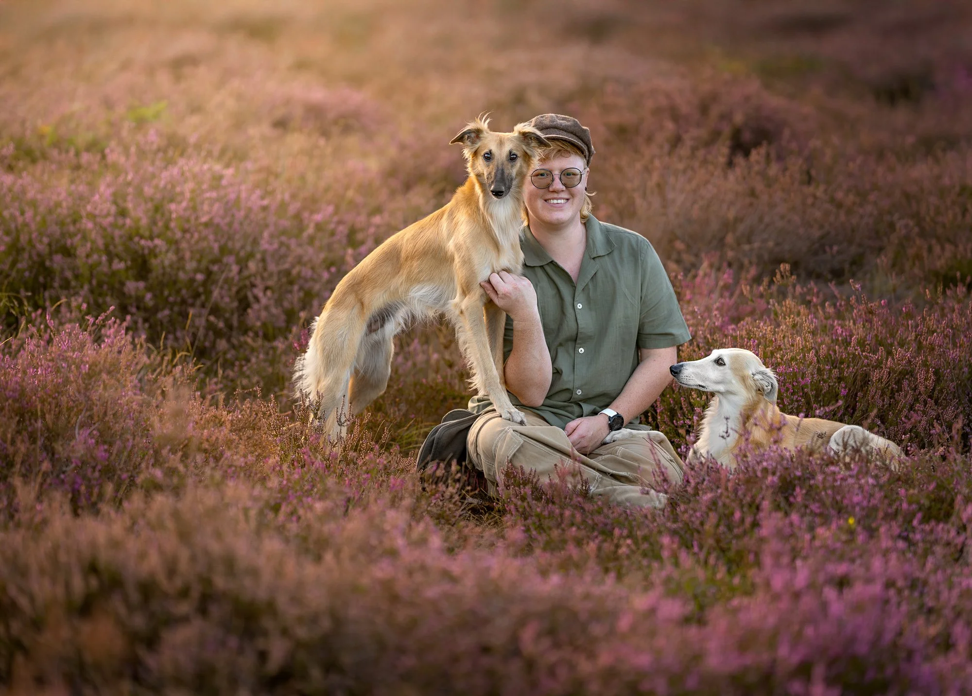 Een persoon met een bril en camouflagebroek zit in een veld met paarse bloemen met twee honden, één staand en één zittend, tijdens zonsondergang.