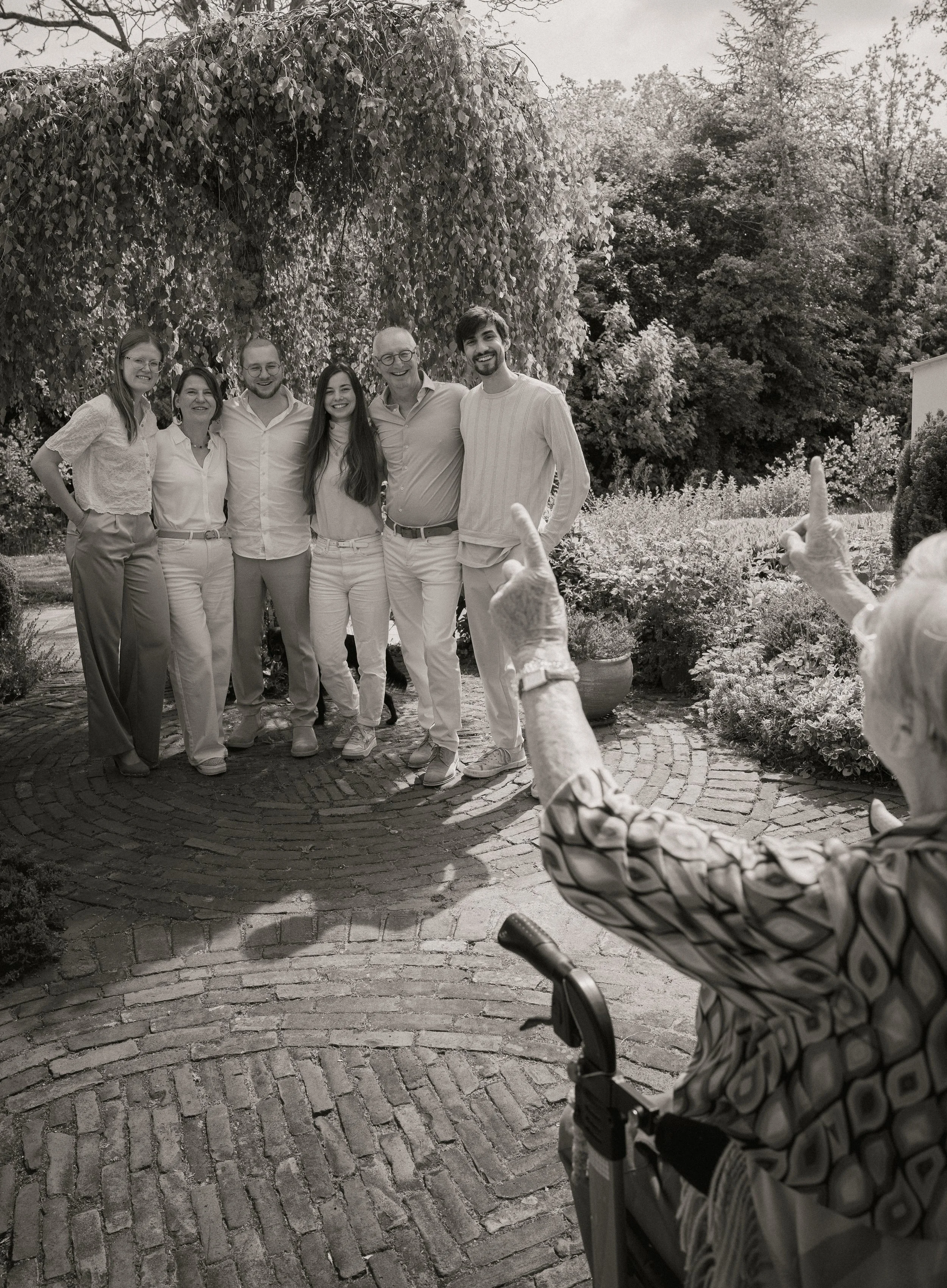Gezicht op een groep mensen die poseert voor een camera buiten, terwijl iemand anders een foto maakt. Er is een kinderwagen in de voorgrond.