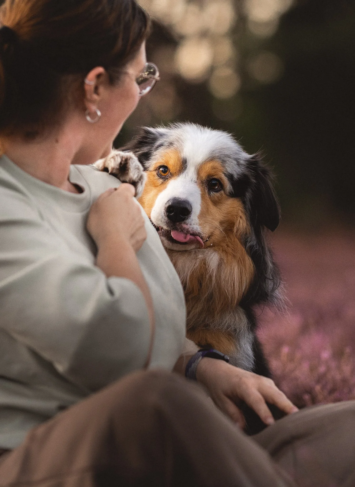 Een persoon met bruin haar en grote oorbellen die een grote Australian Shepherd hond vasthoudt, zowel de persoon als de hond kijken naar de camera, de achtergrond is donkergroen en licht bokeh.