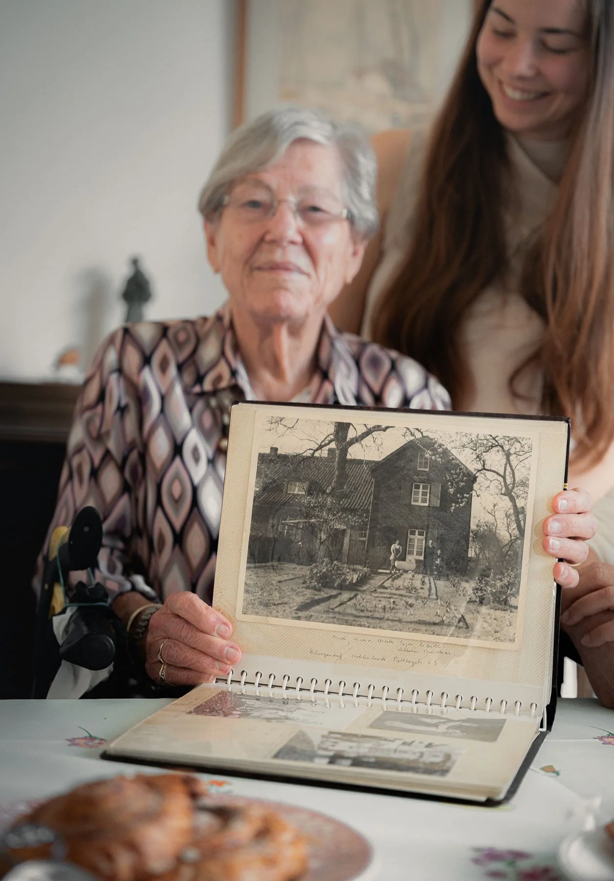 Oudere vrouw met bril toont een zwart-wit foto van een oud huis aan een jonge vrouw met lang bruin haar. Ze zitten samen binnen, op de achtergrond hangt een schilderij.