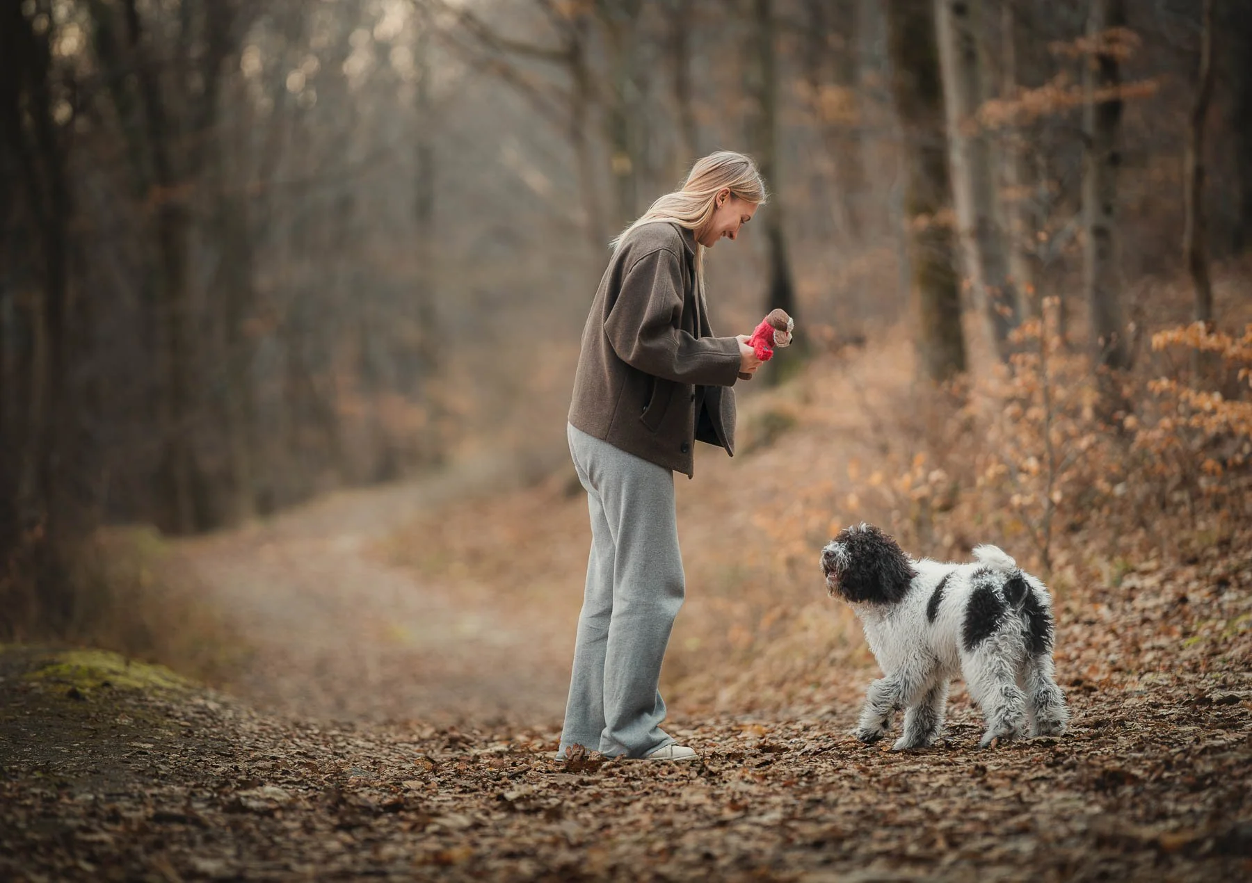 Vrouw met lang blond haar in een grijze joggingbroek en een bruin jack die met haar hond in een bos loopt. Ze lijken te spelen of te communiceren, omringd door herfstbladeren.