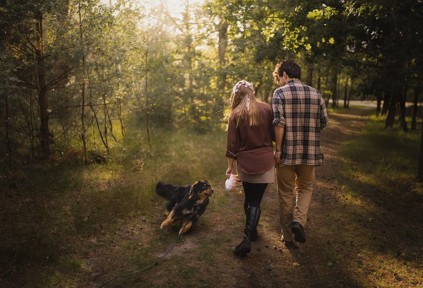 Een stel wandelend door een bos in de middag met een hond, omringd door bomen en zonlicht.
