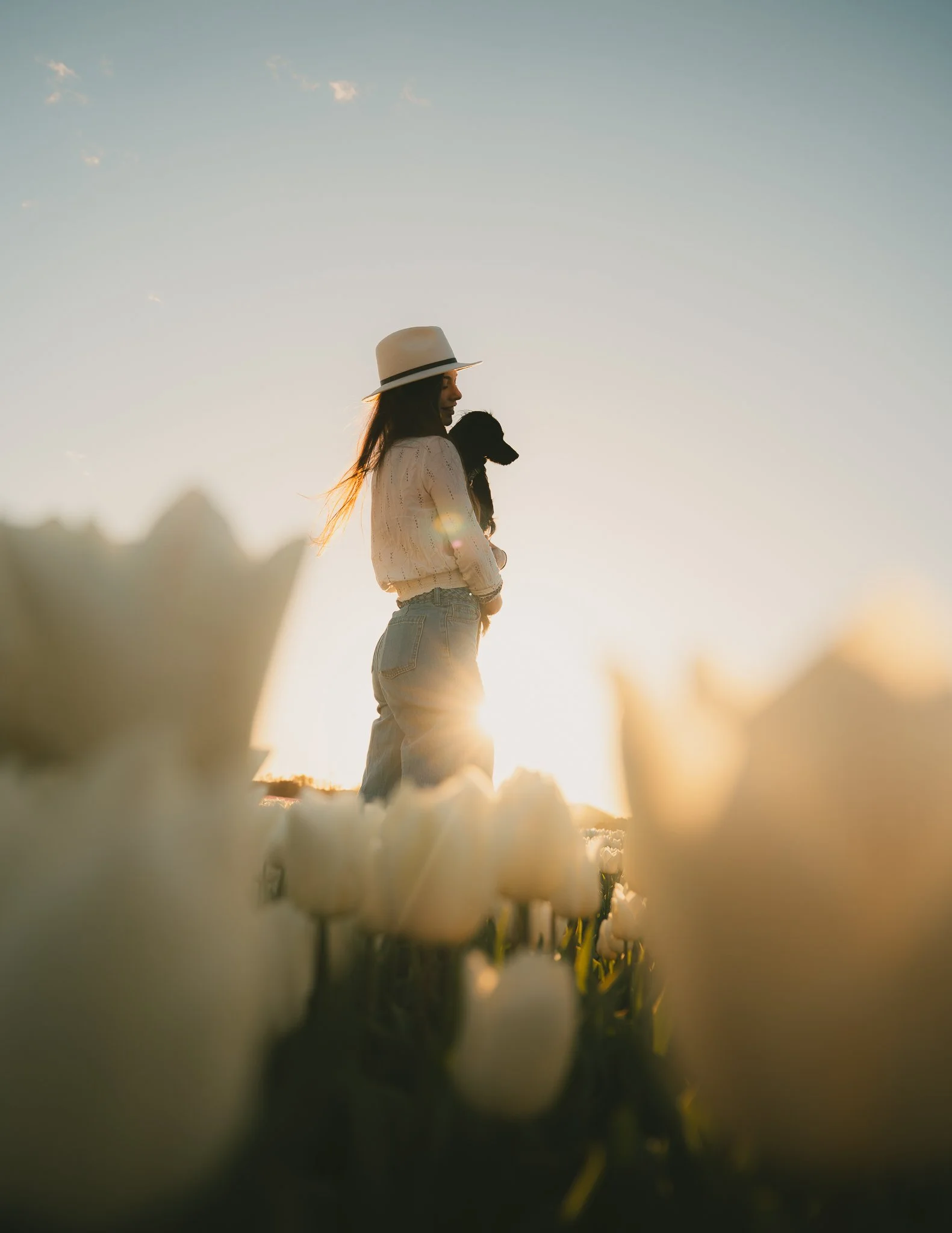 Vrouw met hoed die een hond vasthoudt, zittend in een veld met witte bloemen tijdens zonsondergang.