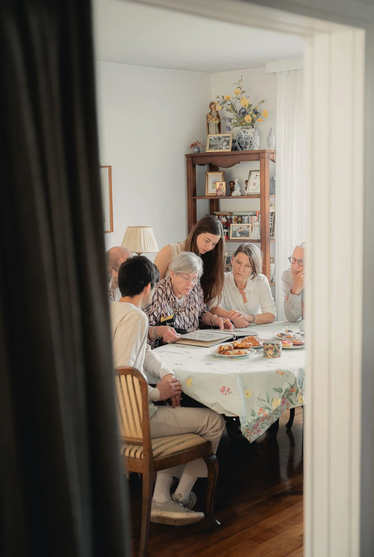 Gezichtspunt op een groep mensen die aan een tafel zitten, met verschillende volwassenen die naar een boek of document kijken. Er staan schalen met gebak op tafel. De foto is genomen door een deurraam, waardoor alleen de rand van de deur zichtbaar is
