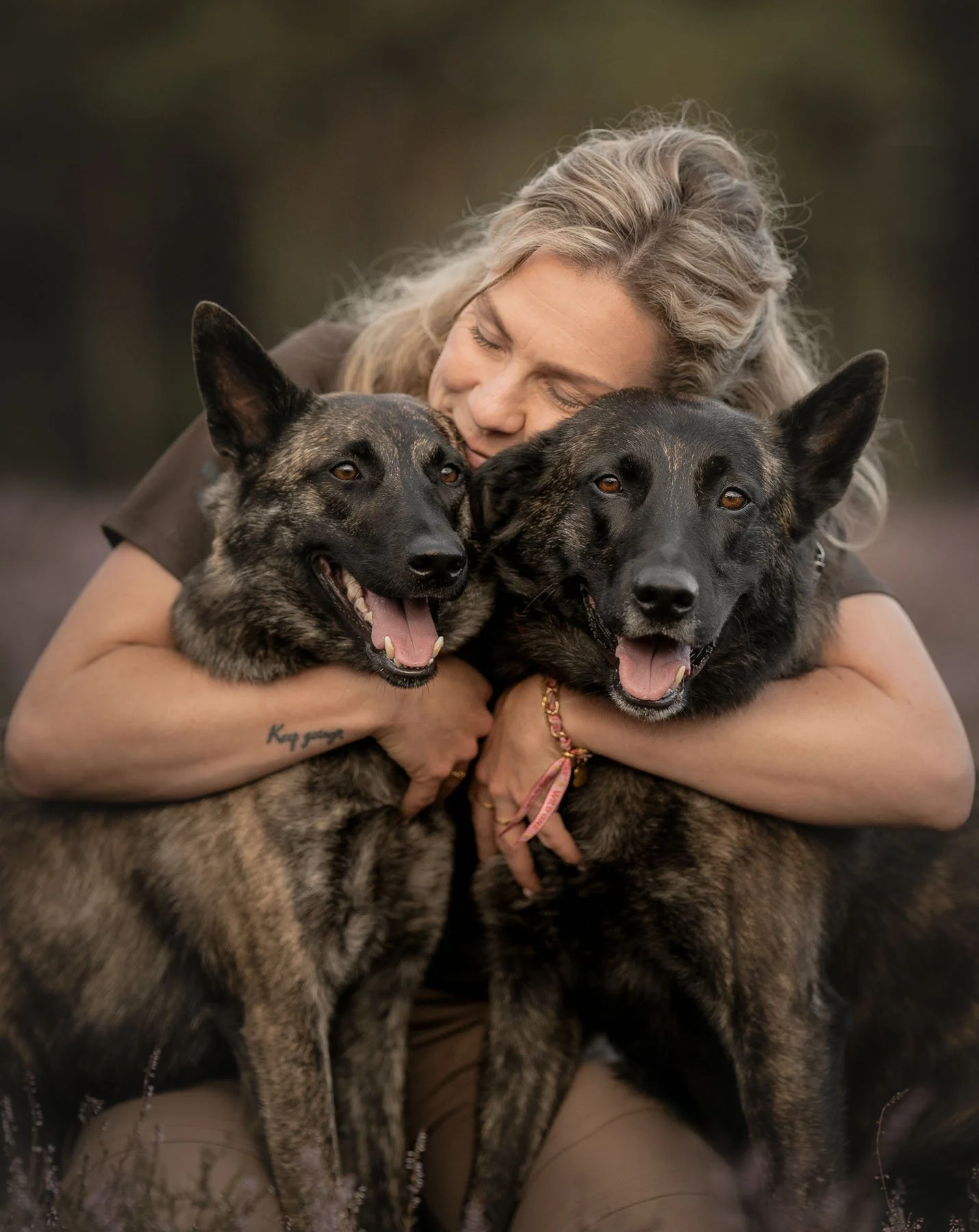Een vrouw die haar ogen sluit en omhelst twee honden met een bruin en zwart bontpatroon.