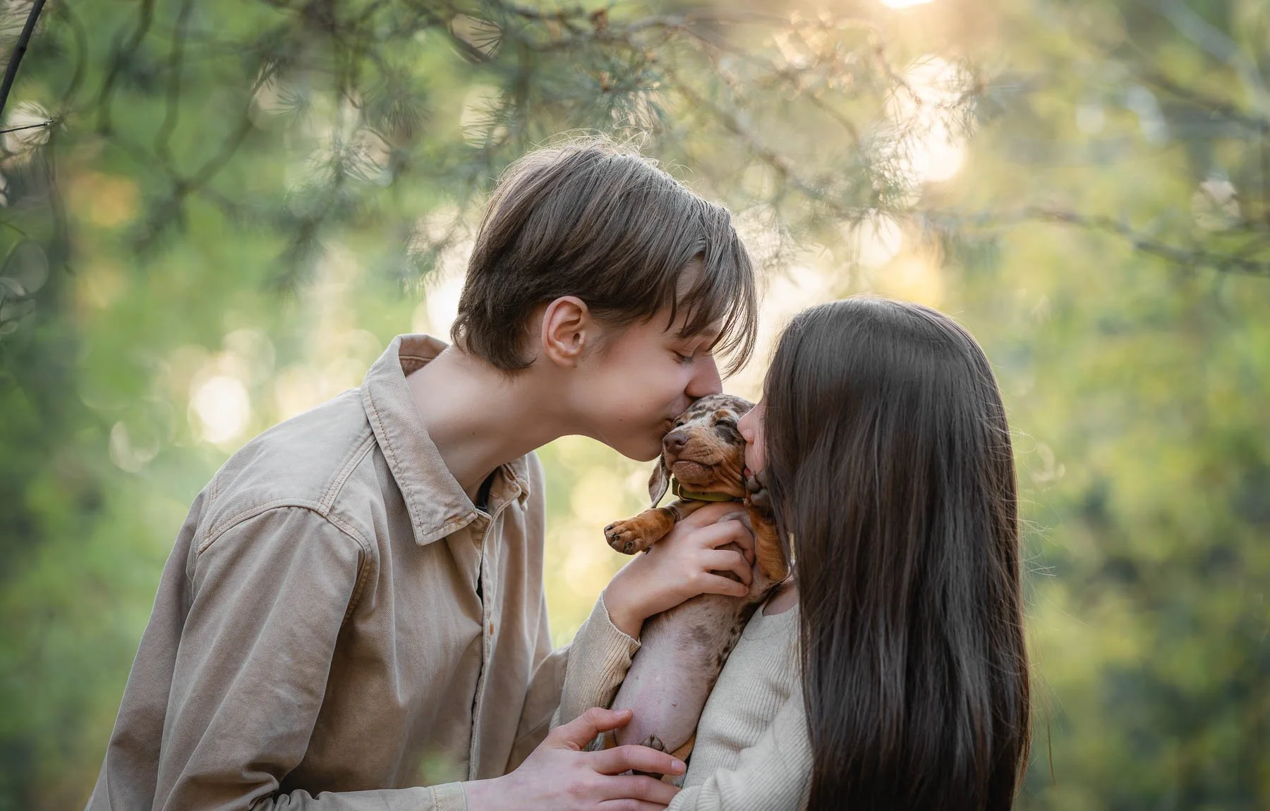 Twee jonge mensen, een man en een vrouw, kussen een puppy met de neus terwijl ze in een bos staan.
