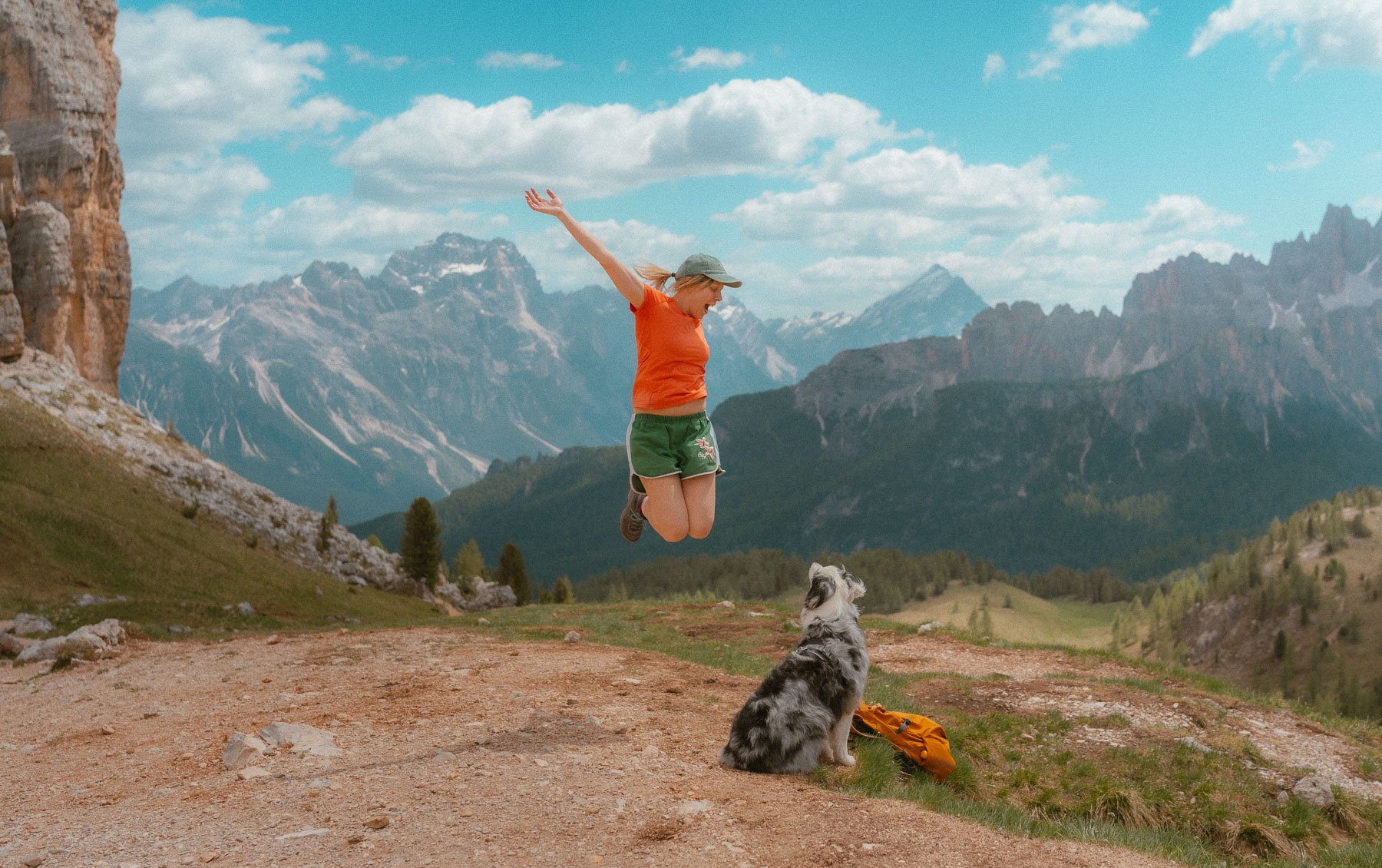 Een vrouw springt in de lucht met haar armen omhoog op een berglandschap, naast een harige hond die op de grond zit en naar haar kijkt, met bergen en wolken op de achtergrond.