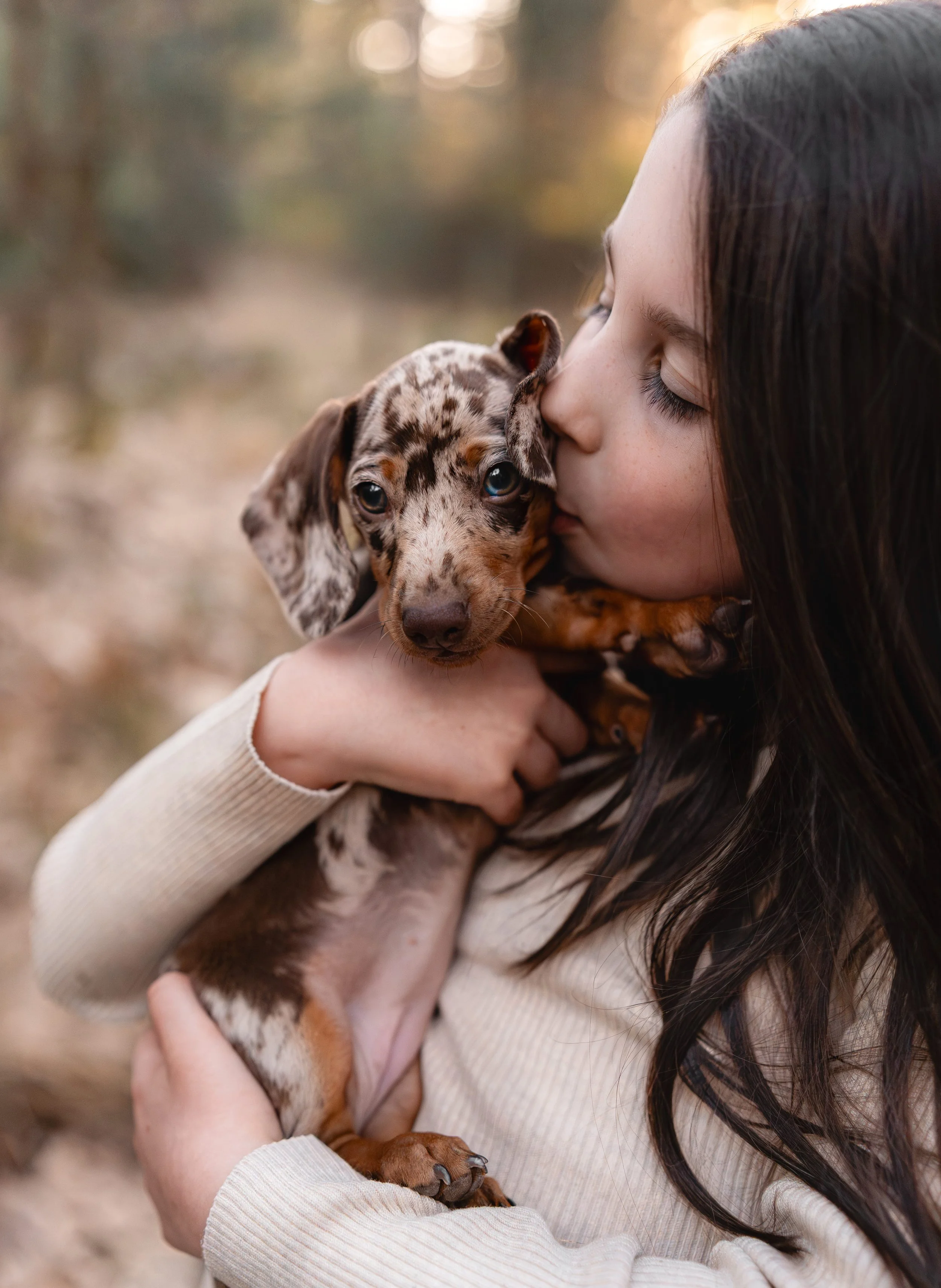 Een meisje dat haar ogen sluit terwijl ze een klein, gevlekte puppy vasthoudt en kusjes geeft in een bosachtige omgeving tijdens zonsondergang.