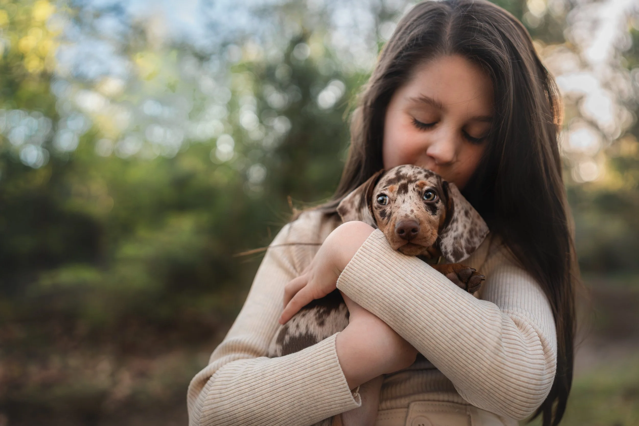 Jonge vrouw met lang bruin haar knuffelt een puppy met een gevlekte vacht in een bosrijke omgeving, met een zachte focus op haar gezicht en het dier.