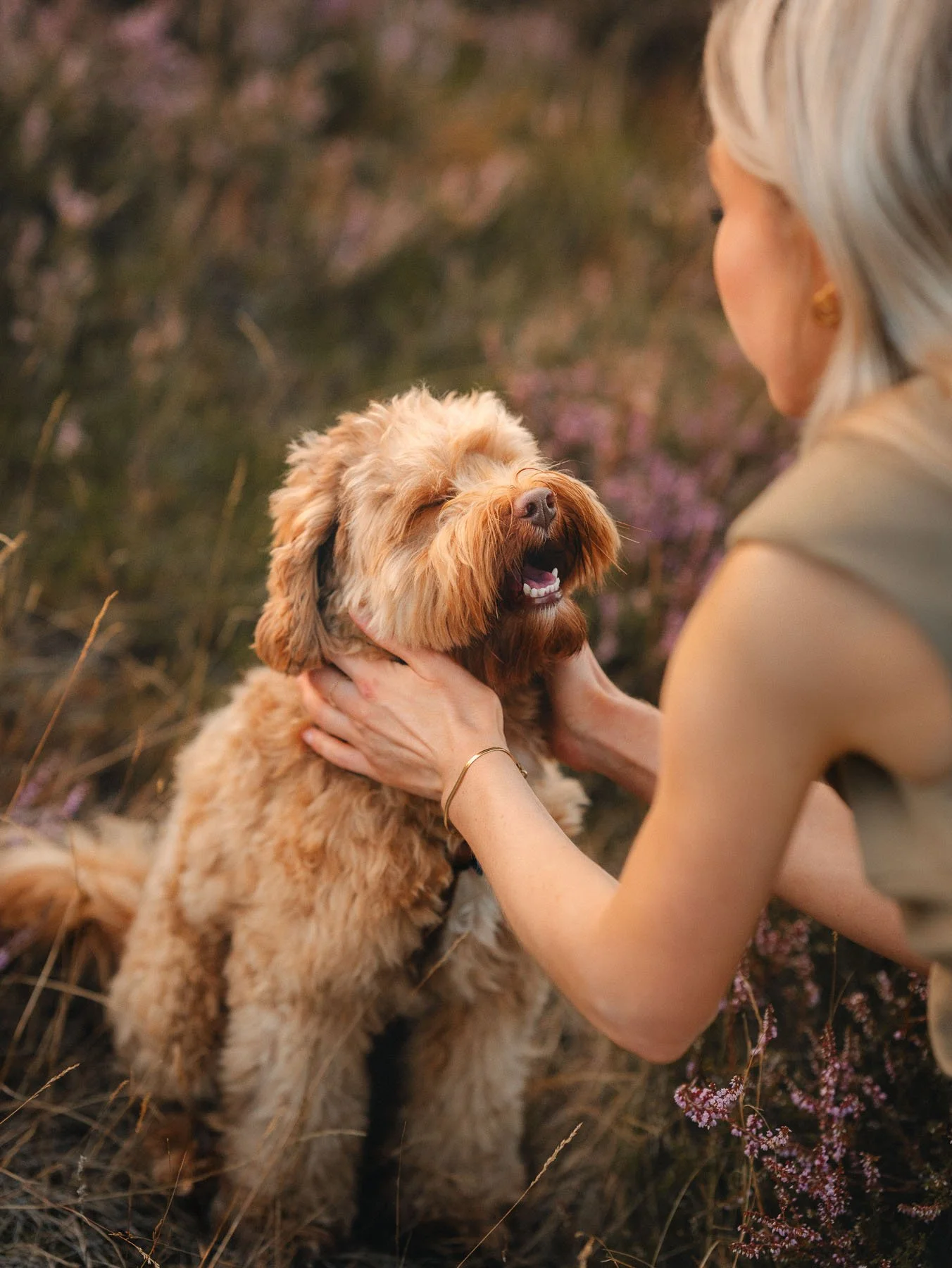 Een vrouw met zilverachtig blond haar knuffelt een vrolijke, lichtebruine, pluizige hond met een brede glimlach in een natuurlijke omgeving met gras en bloemen.