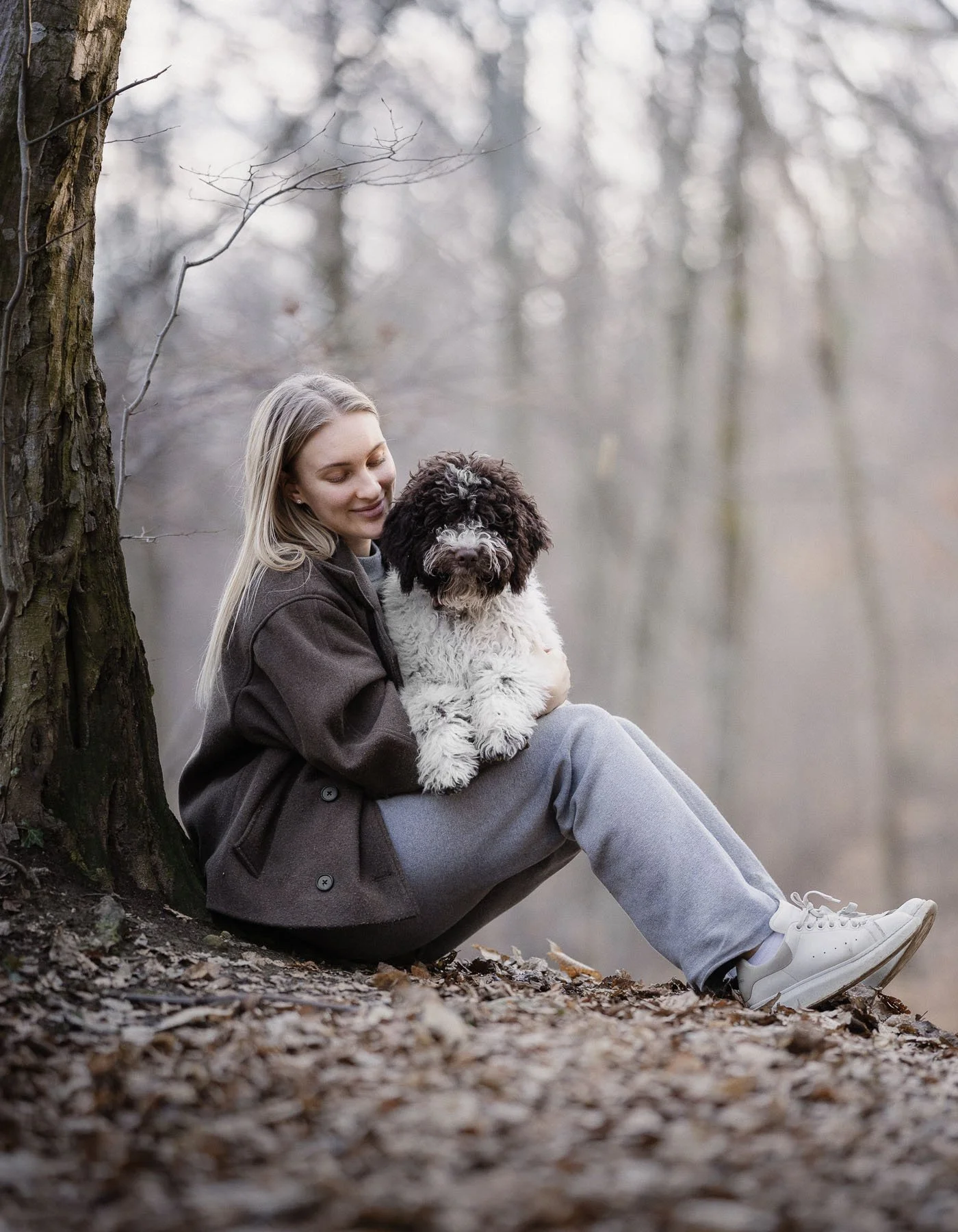 Een jonge vrouw met lang blond haar zit onder een boom in een bos met gevallen bladeren en houdt een grote, knuffelbare zwart-wit gestreepte hond. Ze kijkt liefdevol naar de hond, die op haar schoot ligt.