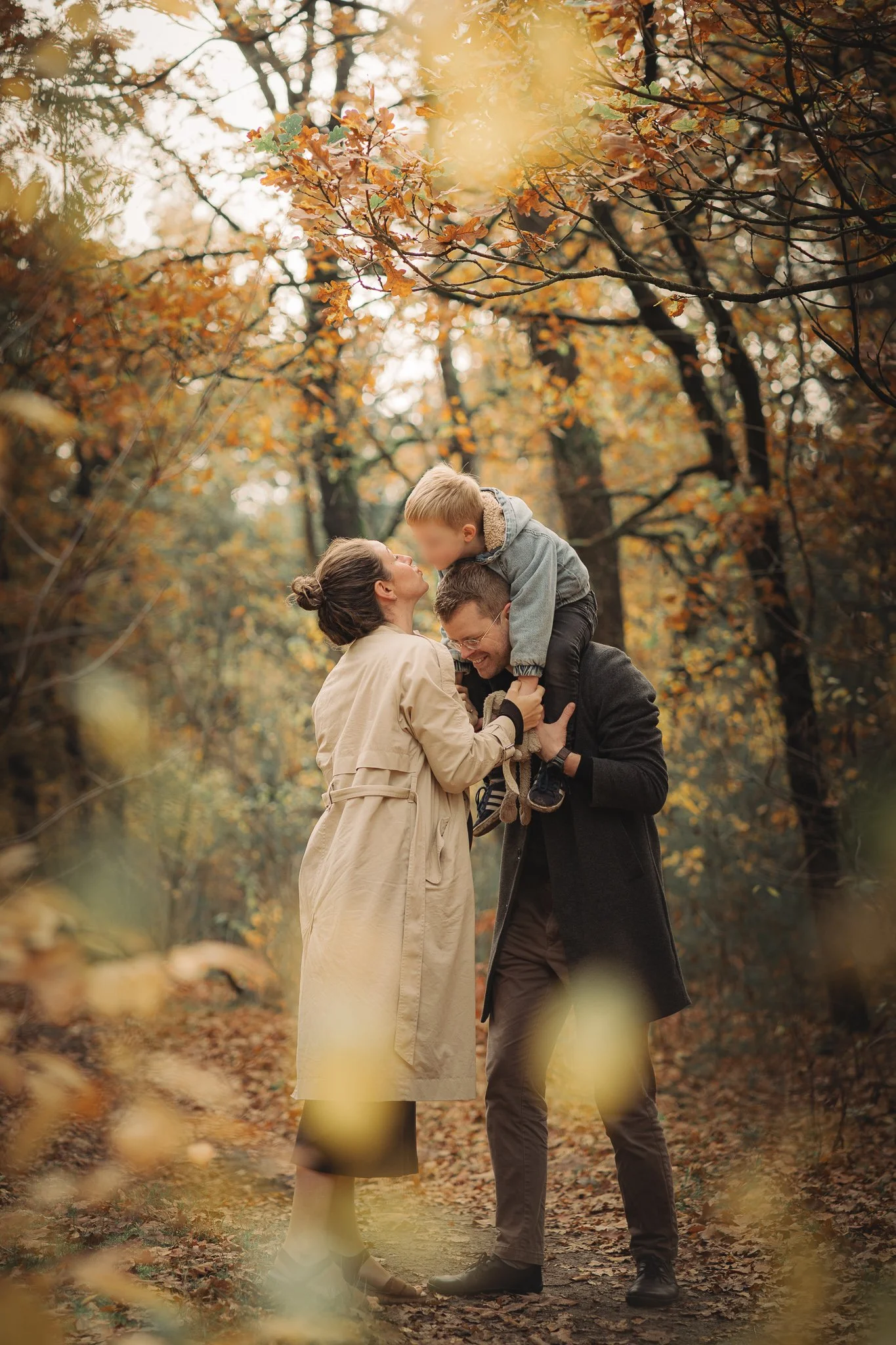 Een vrouw en een man staan in een bos met herfstkleuren. De man tilt een kind op en ze lachen samen.
