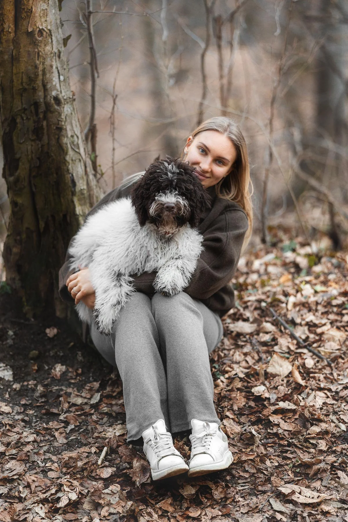 Een jonge vrouw met lang blonde haar zit op de grond omringd door bladeren in een bos, ze houdt een zwart-witte hond in haar armen en knuffelt hem.