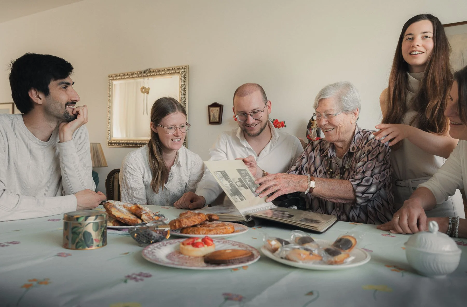 Groep mensen die samen aan een tafel zitten en oude foto's bekijken, omringd door gebak en desserts in een gezellige interieurruimte.