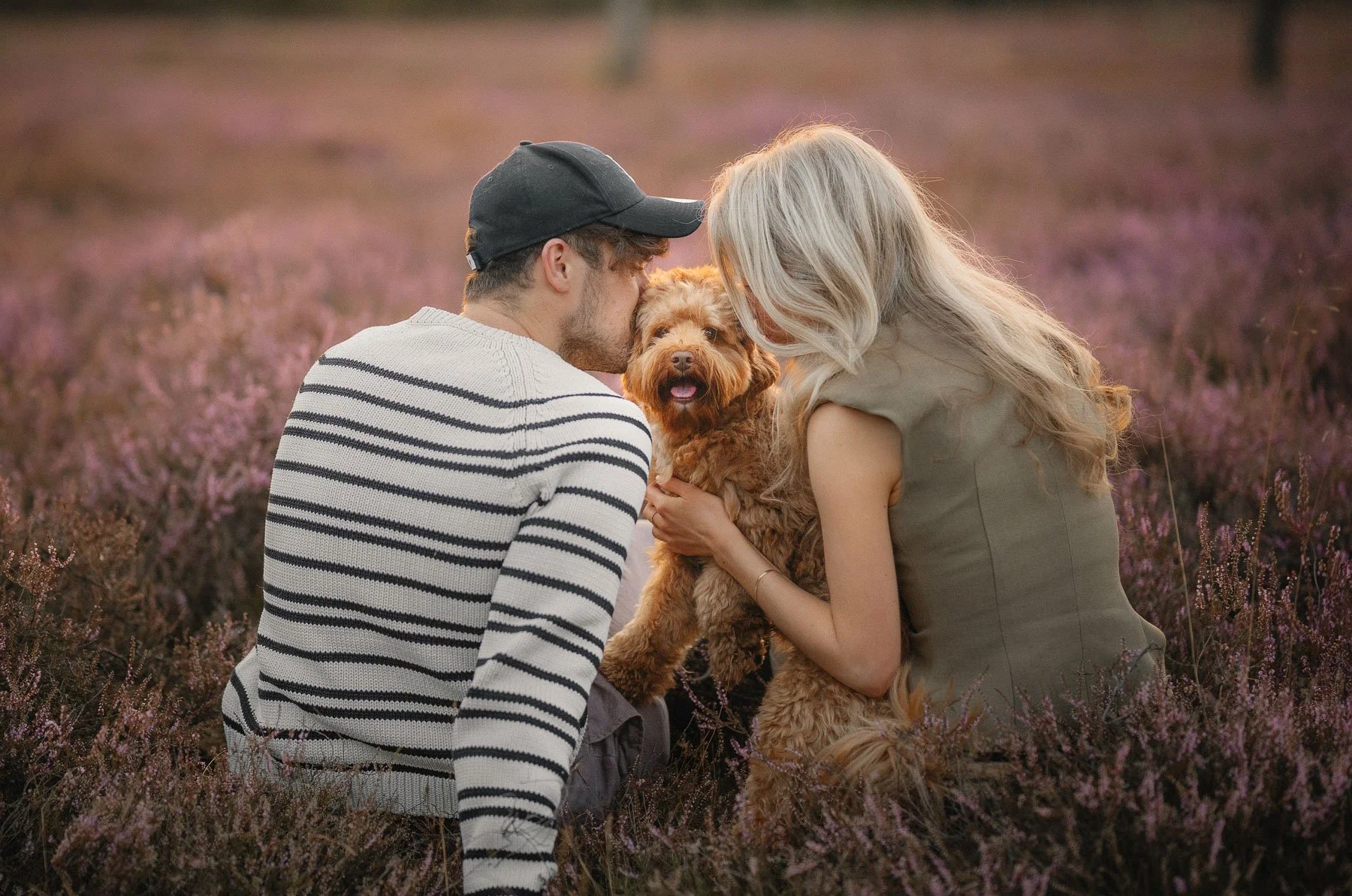 Een man en een vrouw knuffelen een hond in een veld met bloemen bij zonsondergang.