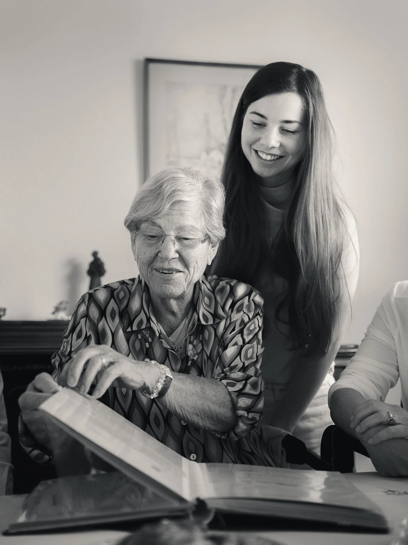 Een oudere man met grijs haar en een kleurrijk overhemd, die binnen een huis is, met een jonge vrouw met lang donker haar die links en achter hem staat, glimlachend. Ze kijken naar een open boek op de tafel.