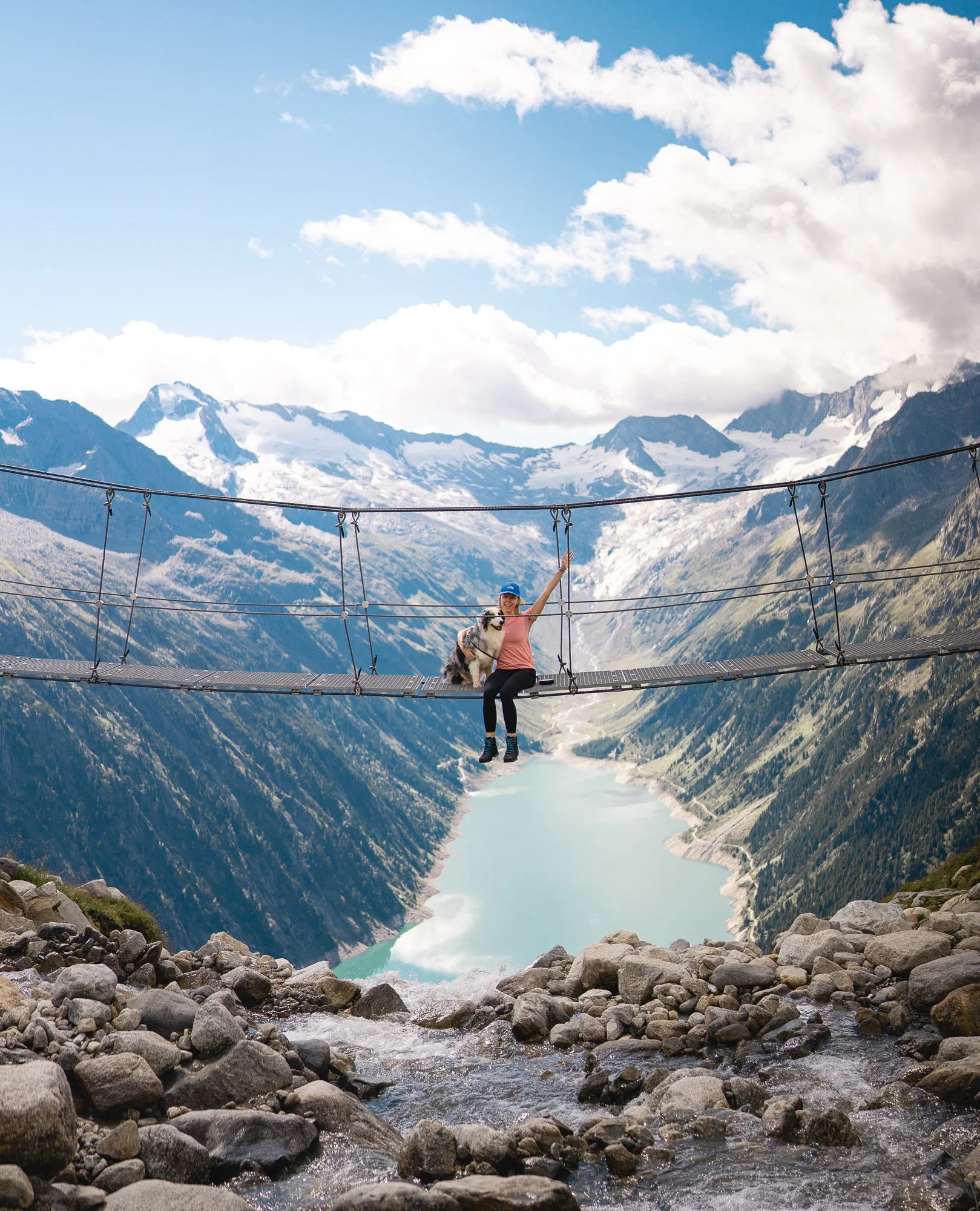 Een vrouw en haar hond steken een hangbrug over hoog boven een rotsachtige rivier, in een bergachtig landschap met besneeuwde bergtoppen en een meer beneden.