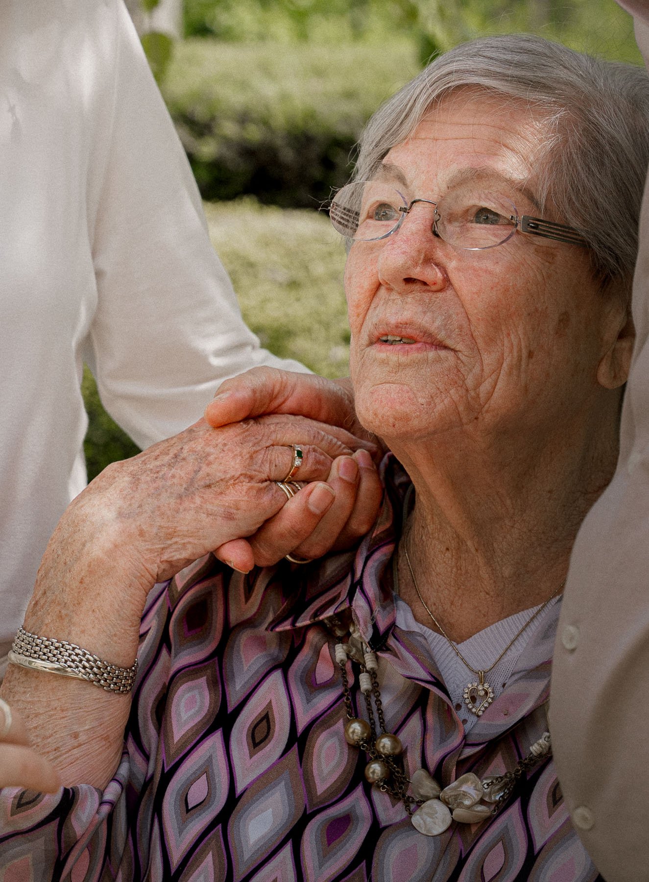 Een oudere vrouw met grijs haar en bril, die op haar kin wordt vastgehouden door iemand anders. Ze draagt een parelsnoer en een ketting, en heeft een ring op haar vinger. Ze zit buiten op gras, gekleed in een kleurrijk, geometrisch patroon blouse.