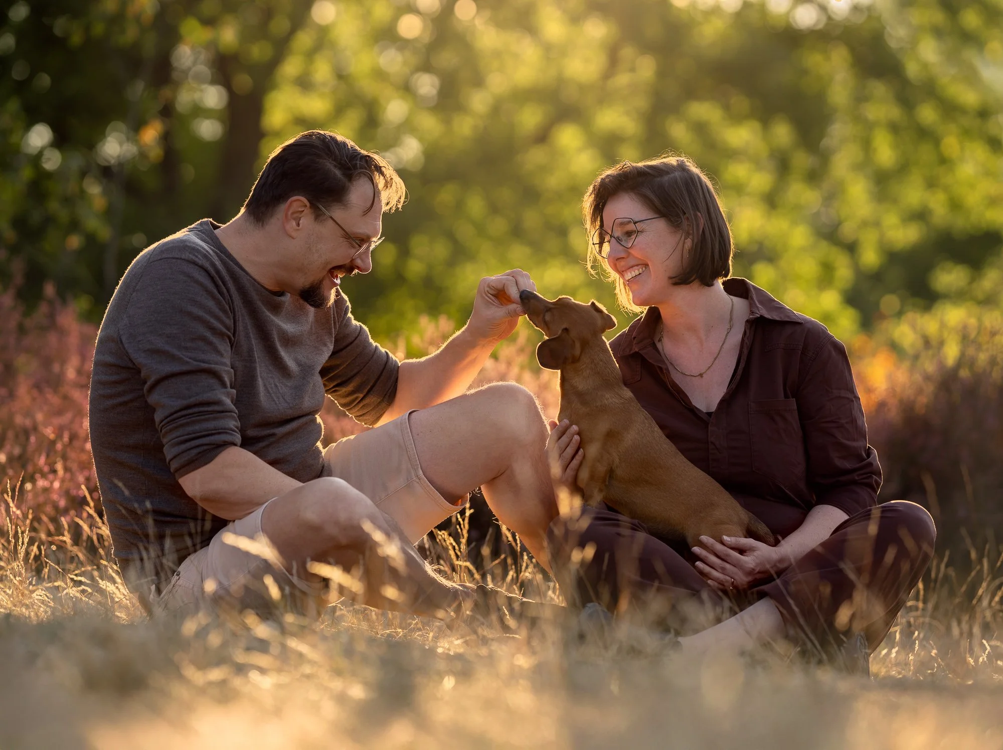 Intieme fotoshoot van teckel en twee eigenaren in de heide met focus op connectie en natuur.