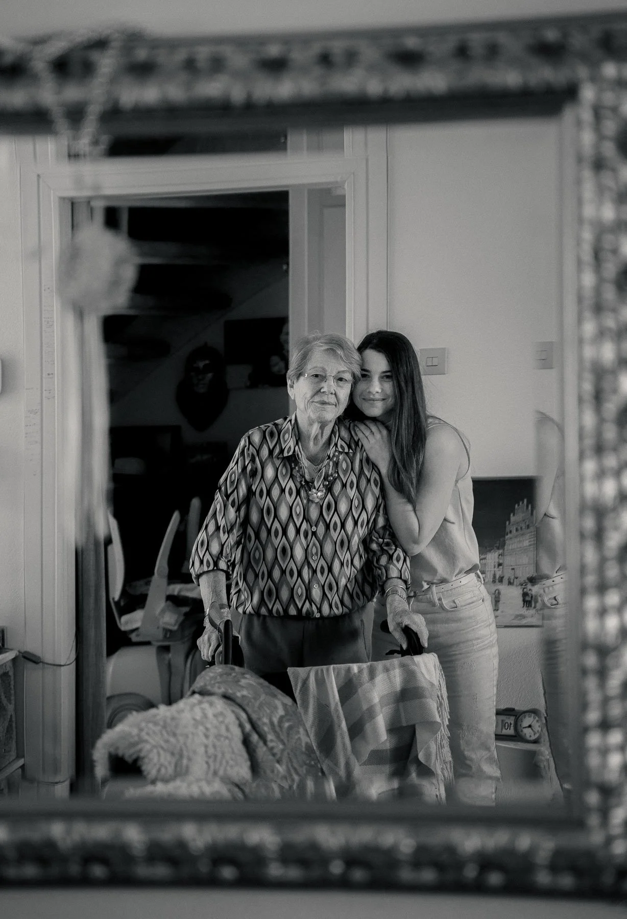 Een oudere vrouw en een jonge vrouw die samen in de spiegel naar de camera kijken, met de oudere vrouw die ondersteunt met een wandelstok.