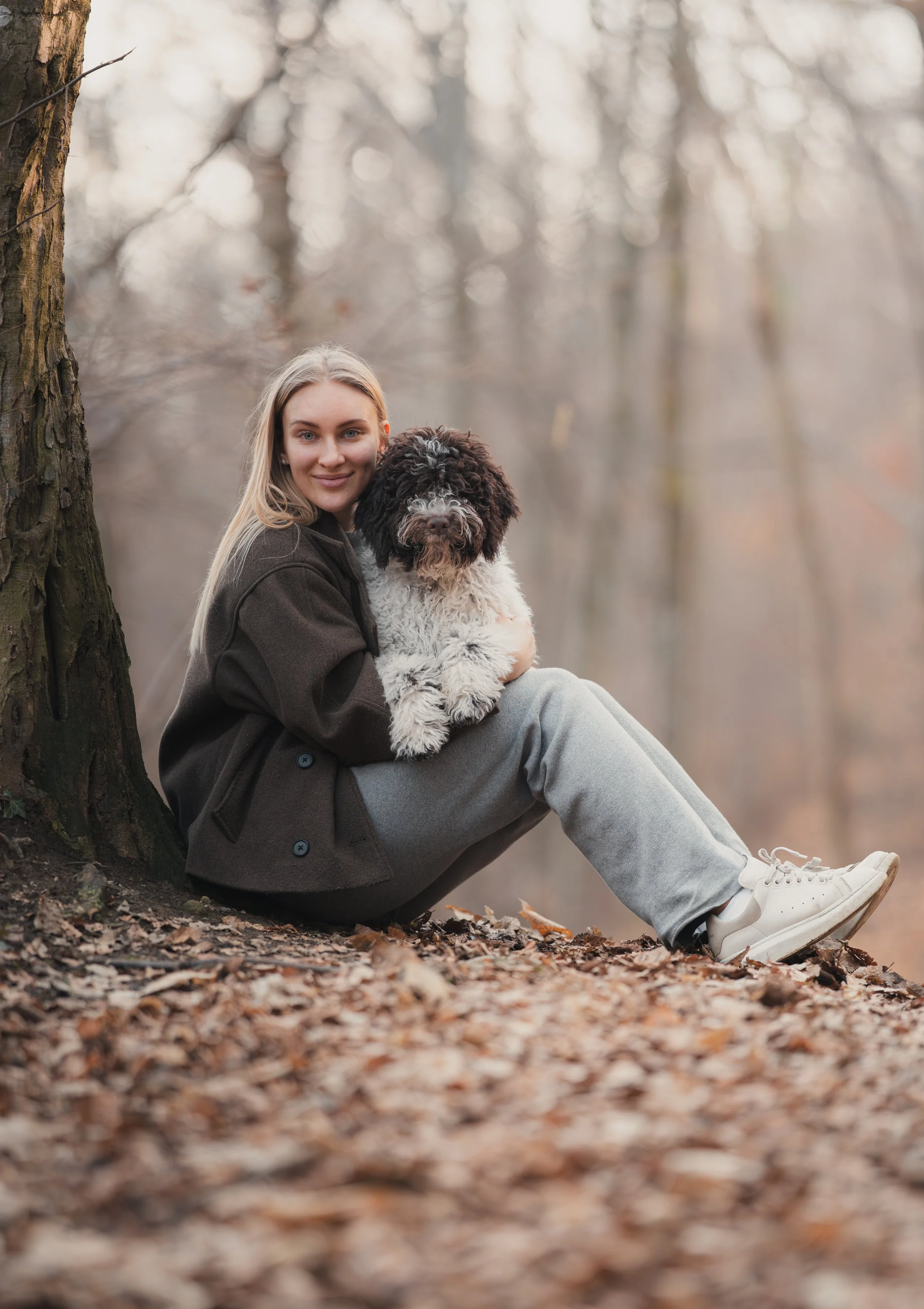Vrouw die op een boomstronk in een bos zit, haar hond vasthoudt, herfstachtig landschap met bladeren op de grond en een schommel in de achtergrond.