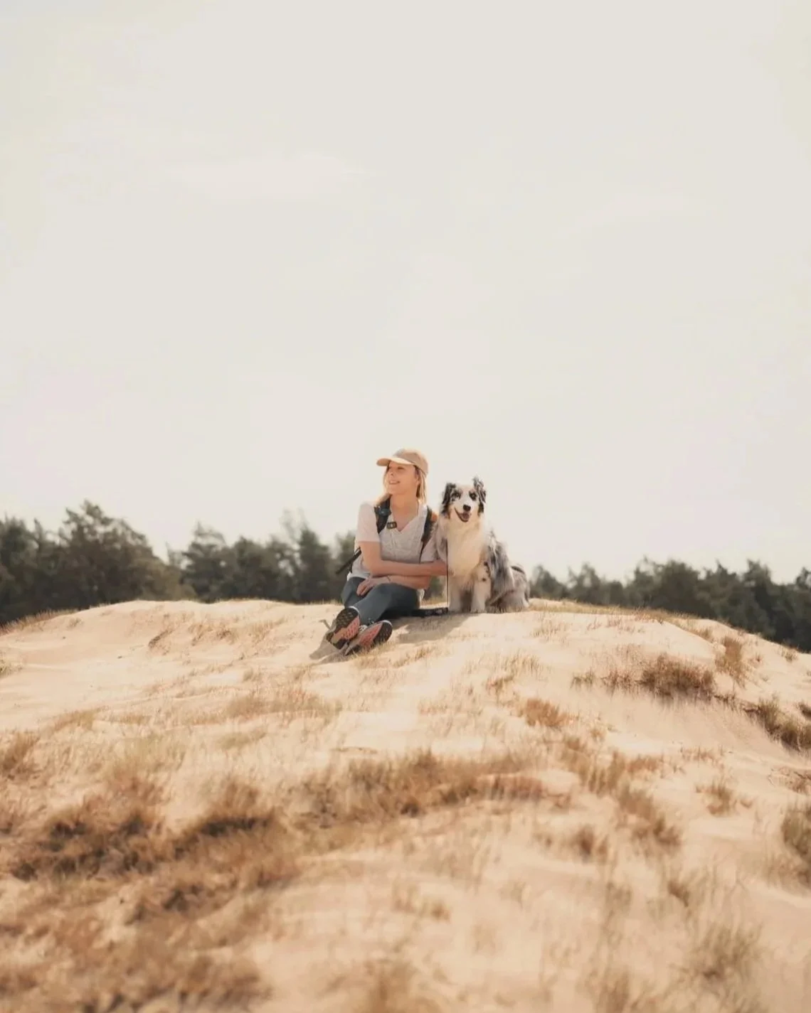 Een vrouw zit op een zandheuvel met haar Australian Shepherd hond, in een open veld met bomen op de achtergrond, op een zonnige dag.
