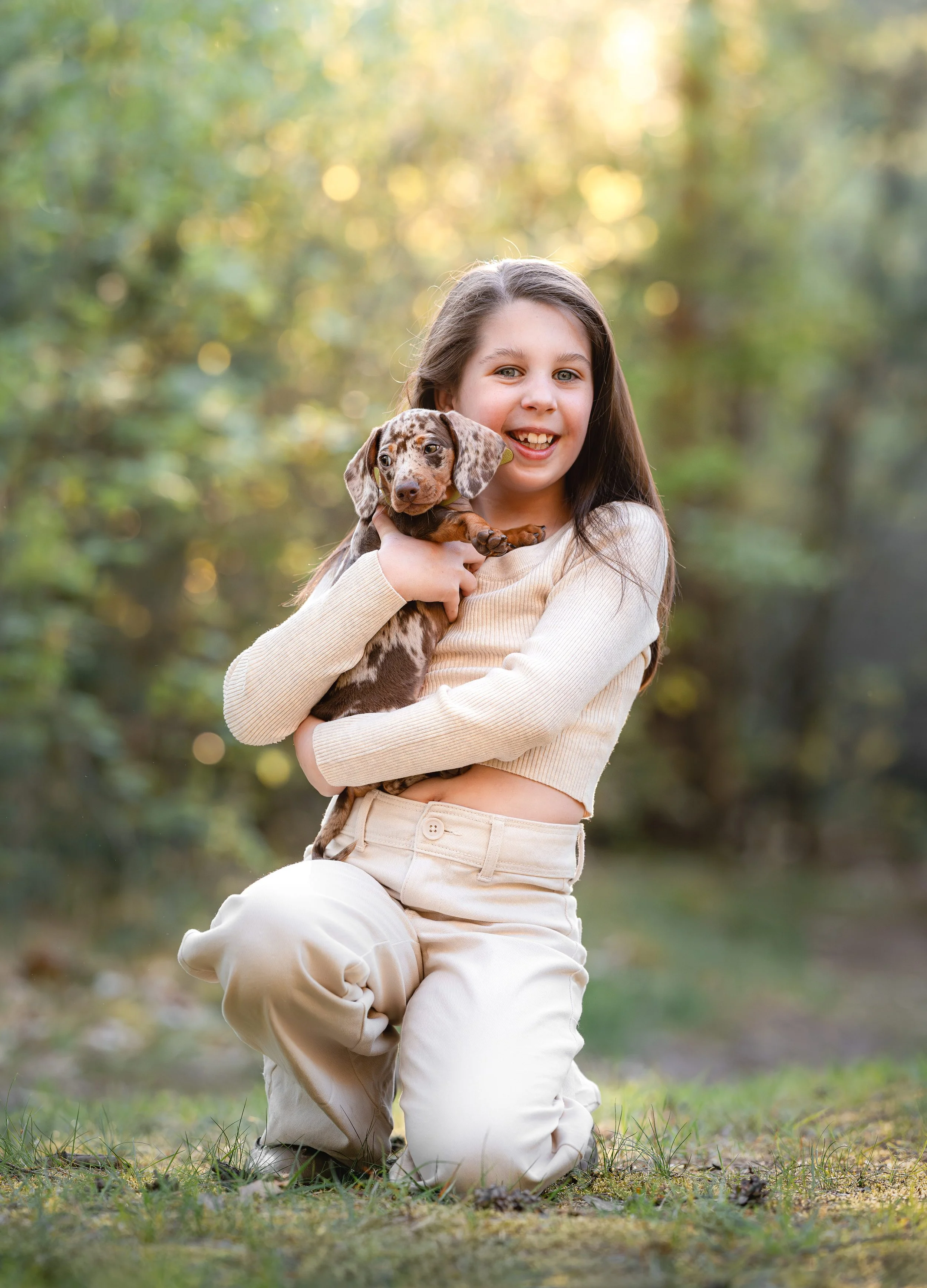 Jonge vrouw die op de knieën zit in een park met groene bomen op de achtergrond en een hond vasthoudt.