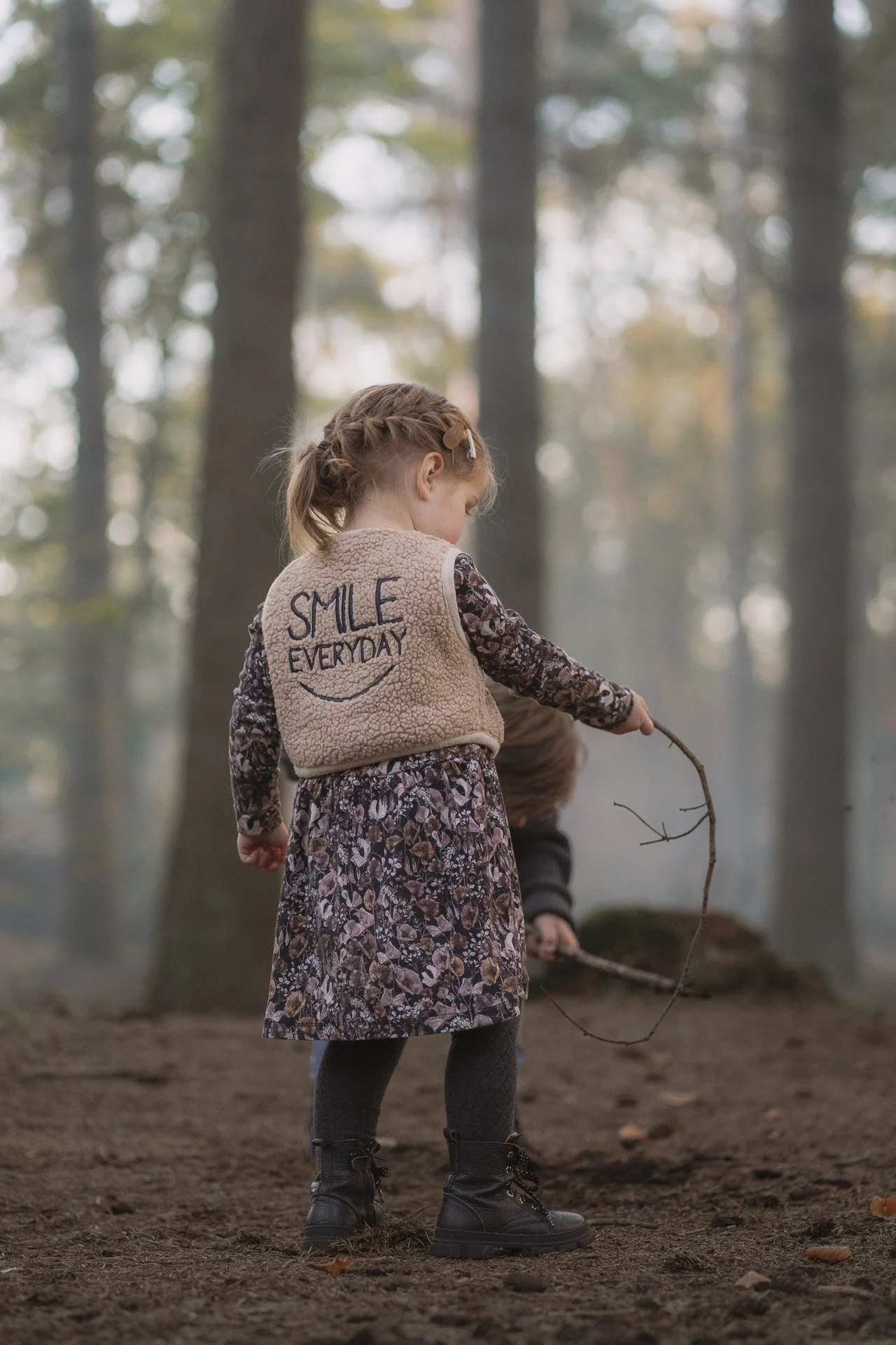 Een jonge meid met een geruite jurk, een beige vest met de tekst 'Smile Everyday' en een glimlach tekening, staat in een bos en houdt een tak in haar hand.