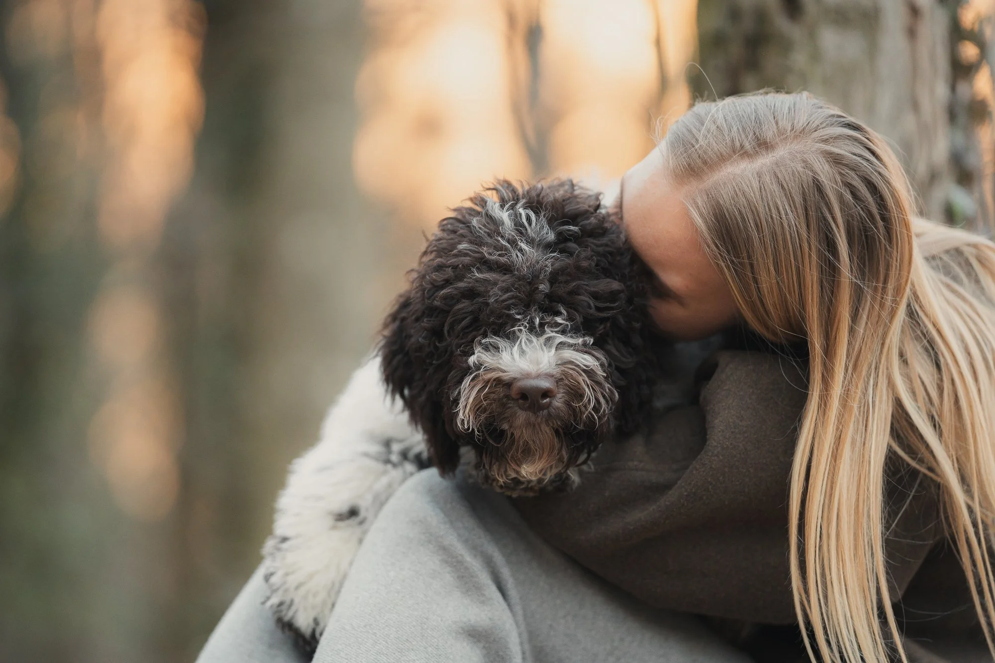 Een persoon met lang blond haar knuffelt een zwart-witte krulharige hond buiten tijdens zonsondergang.