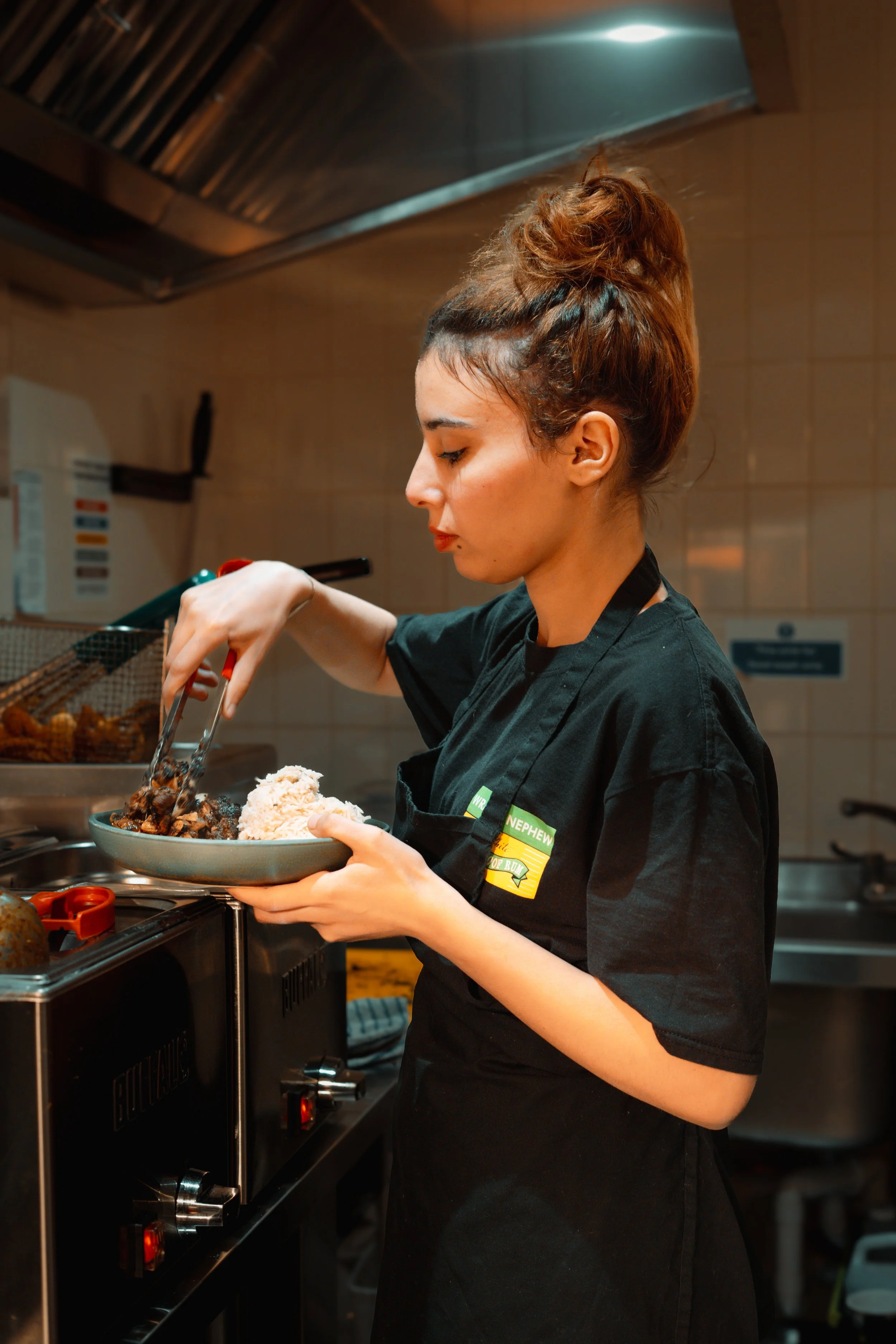 A young woman in a black chef's coat serving food from a pan onto a plate in a professional kitchen.