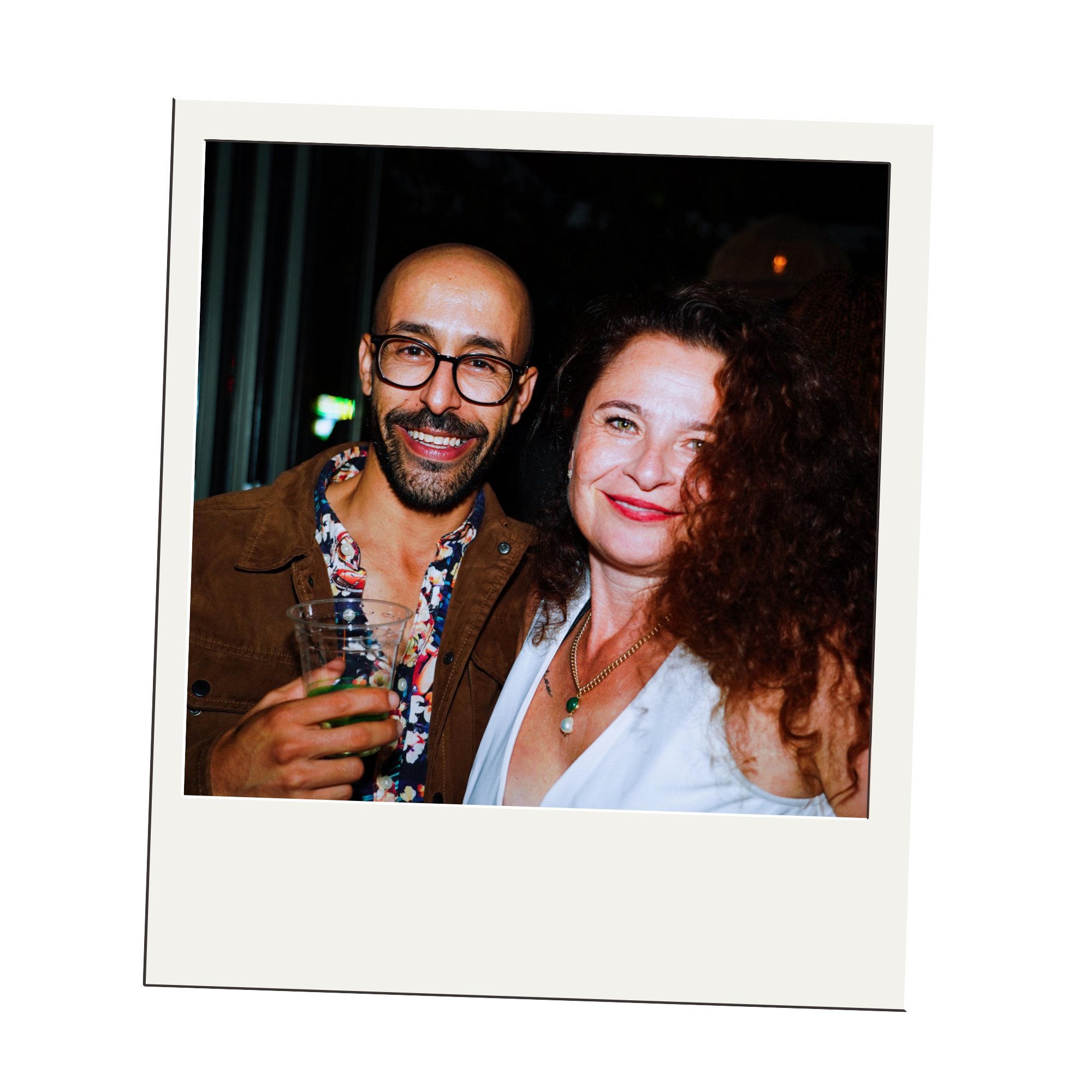 A smiling man with glasses and a beard holding a drink standing next to a smiling woman with curly hair, both at a social event.
