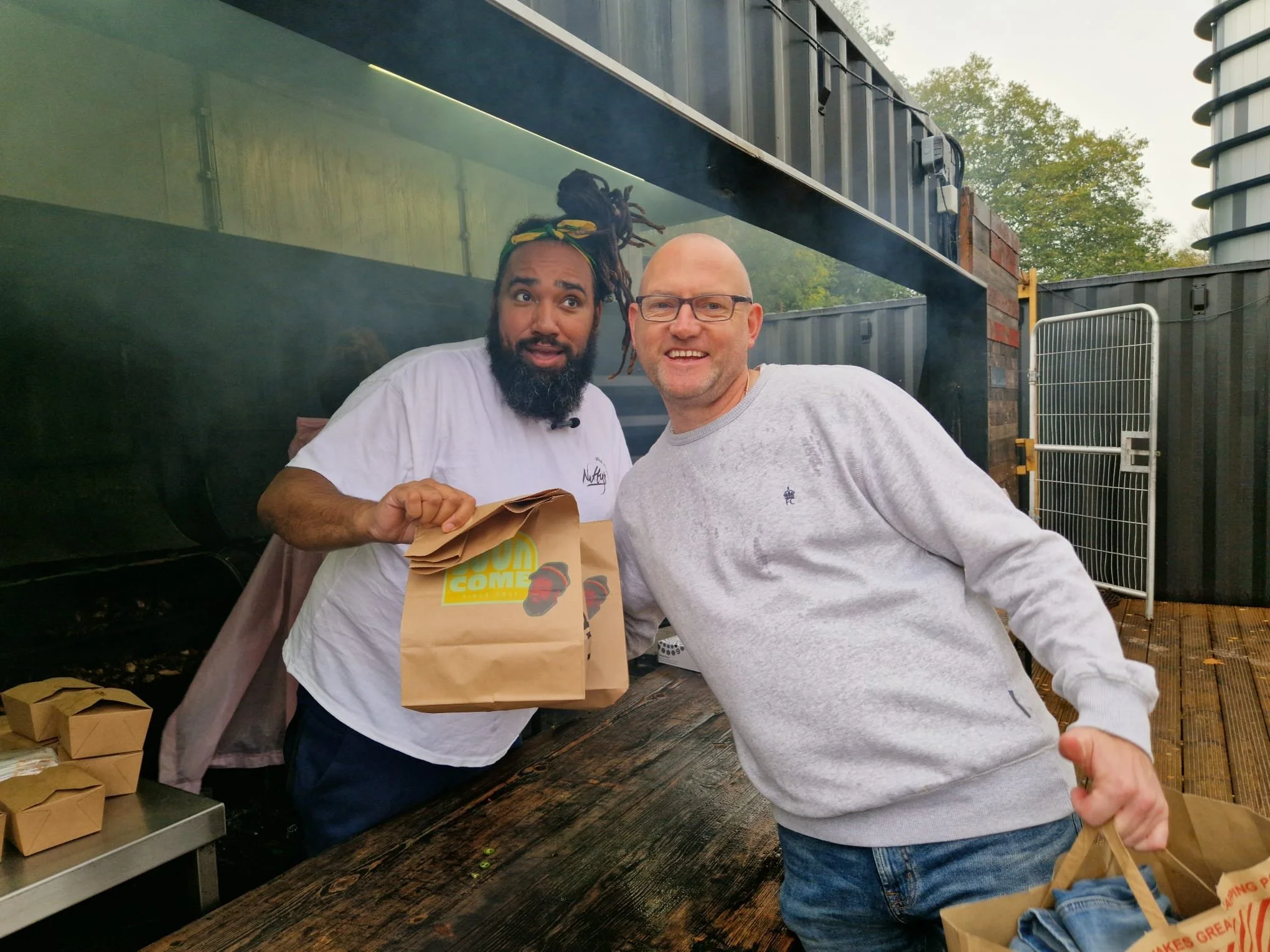 Two men smiling at an outdoor food stand, one holding a paper bag. The man on the left has dreadlocks and a beard, wearing a white t-shirt. The man on the right is balding with glasses, wearing a light gray sweatshirt. There are food boxes on the table, and trees in the background.