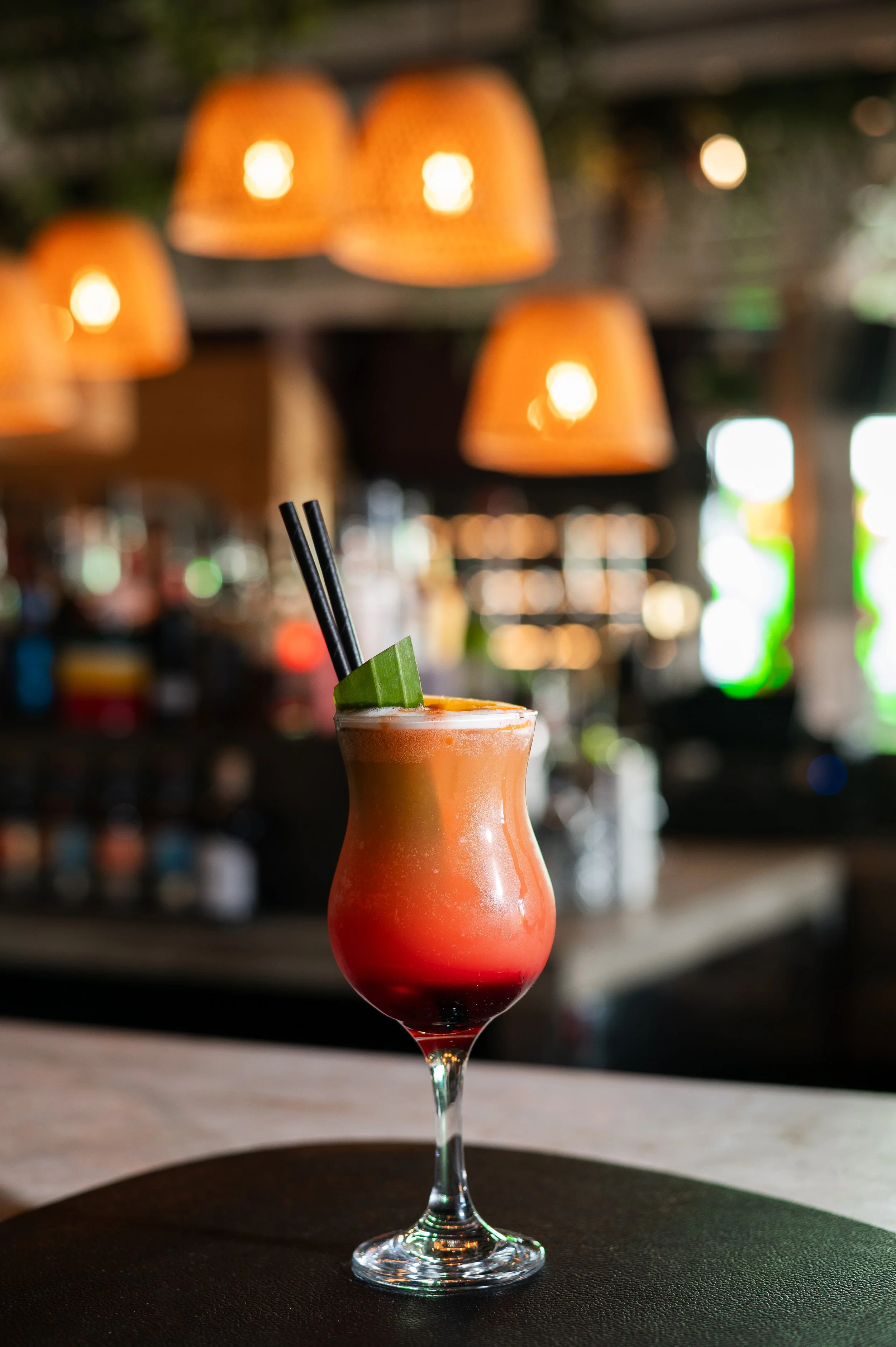 Colorful cocktail in a hurricane glass garnished with lime and black straws, seated on a bar counter with warm orange hanging lights in the background.