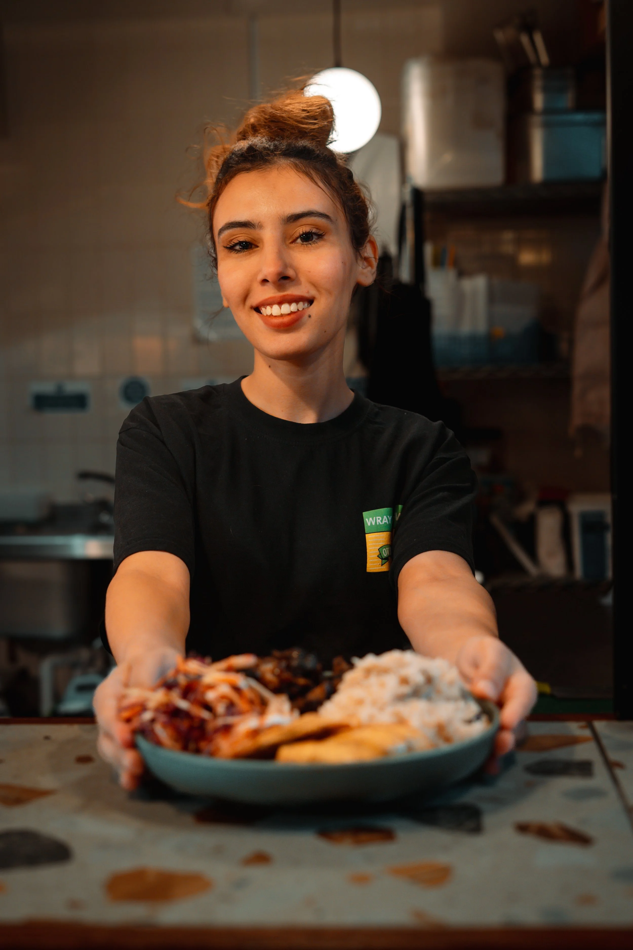 A woman with a bun hairstyle and black T-shirt serving a plate of food at a restaurant counter.