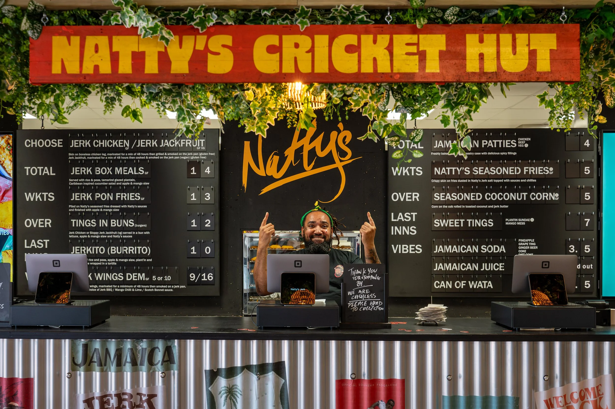 A man with dreadlocks and a beard behind a counter at Naty's Cricket Hut. The shop offers various Jamaican foods and drinks, with colorful signs listing menu items. The storefront is decorated with green foliage and a prominent red and yellow sign reading 'Natt y's Cricket Hut'.