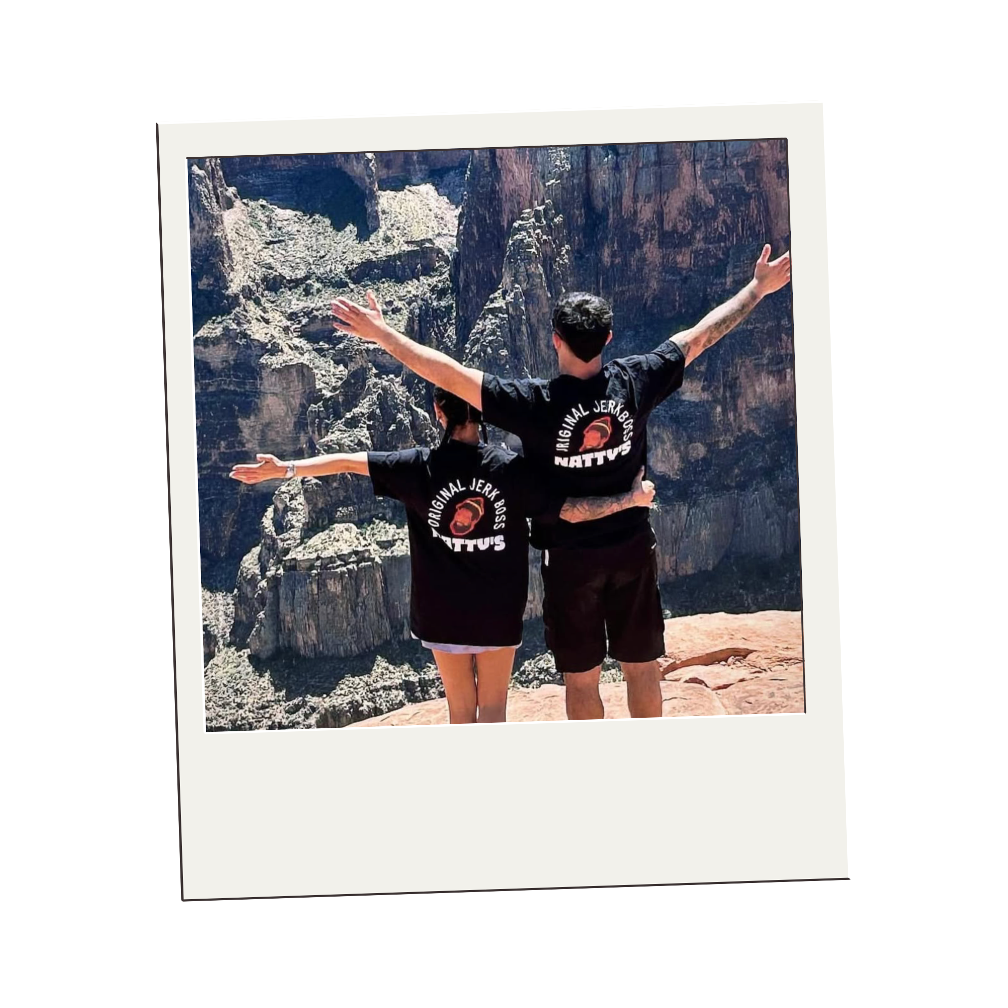 Two people wearing matching black t-shirts with a logo and text, standing at the edge of a canyon with arms outstretched, overlooking rocky cliffs.
