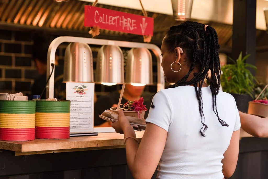 A woman ordering food at a counter with a sign that says 'Collect Here,' in a restaurant or food establishment.