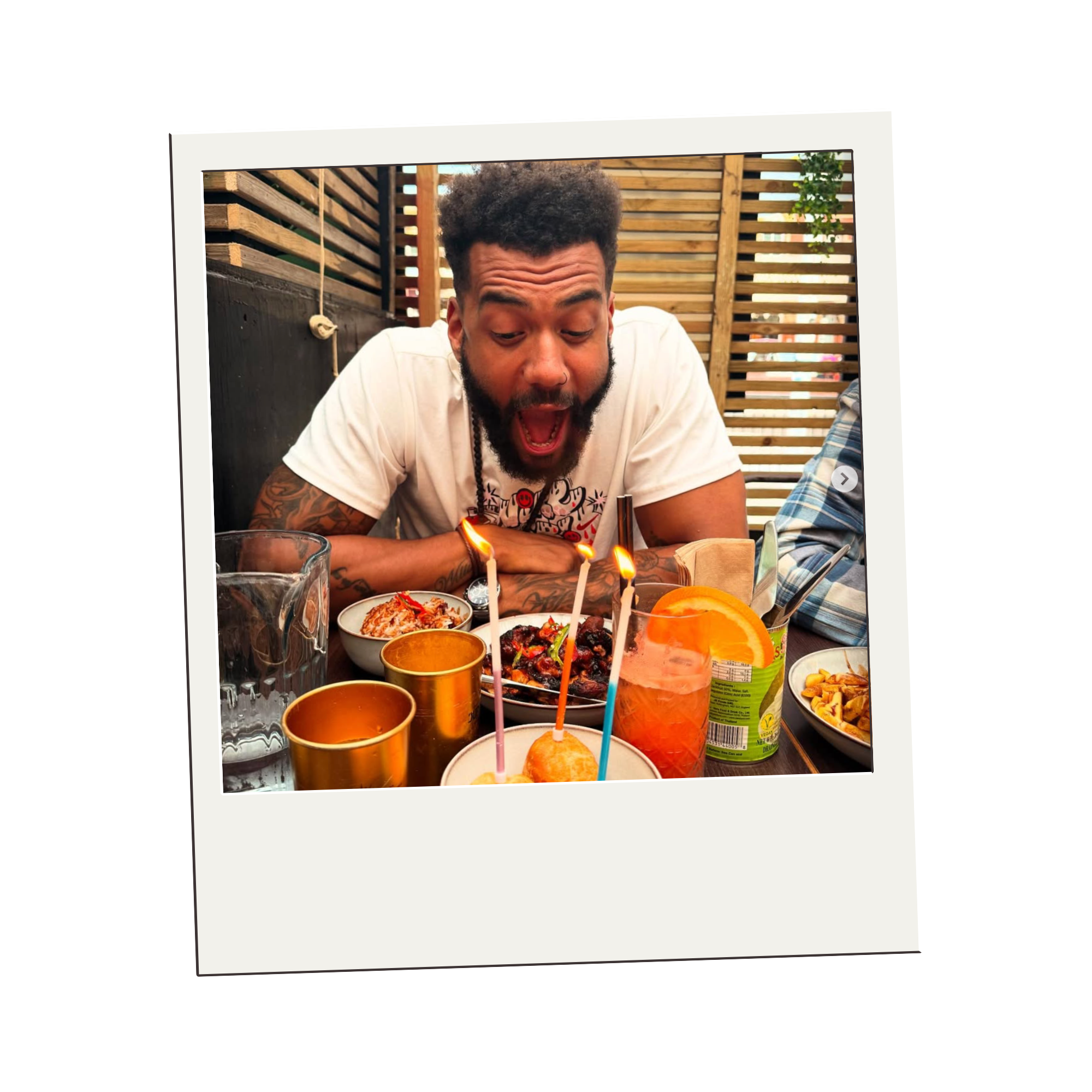 A man celebrating a birthday with lit candles on a cake, surrounded by snacks and drinks on a table.