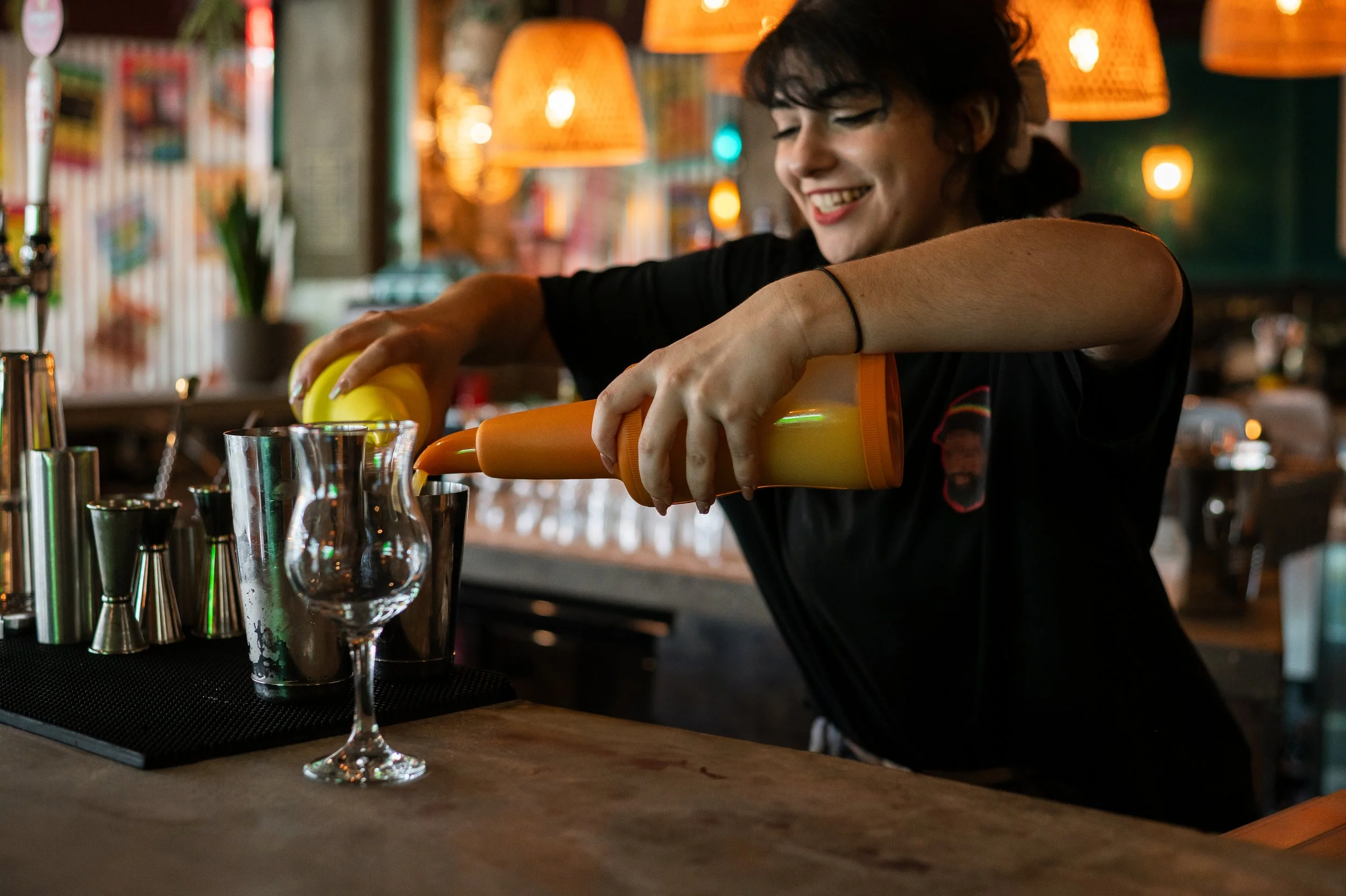 A woman smiling while pouring orange liquid from a squeeze bottle into a glass at a bar or restaurant, with bar tools and glasses on the counter.