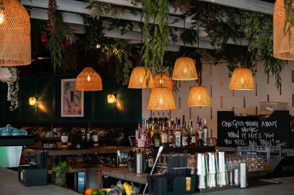 Interior of a bar with hanging woven rattan pendant lights, greenery on the ceiling, bottles of alcohol on the bar counter, and a chalkboard sign with a quote about music and feelings.