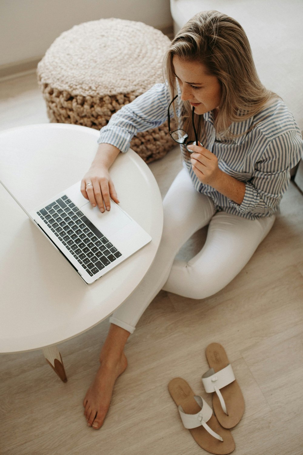 Woman in striped shirt using laptop on white table, holding glasses, with sandals on floor